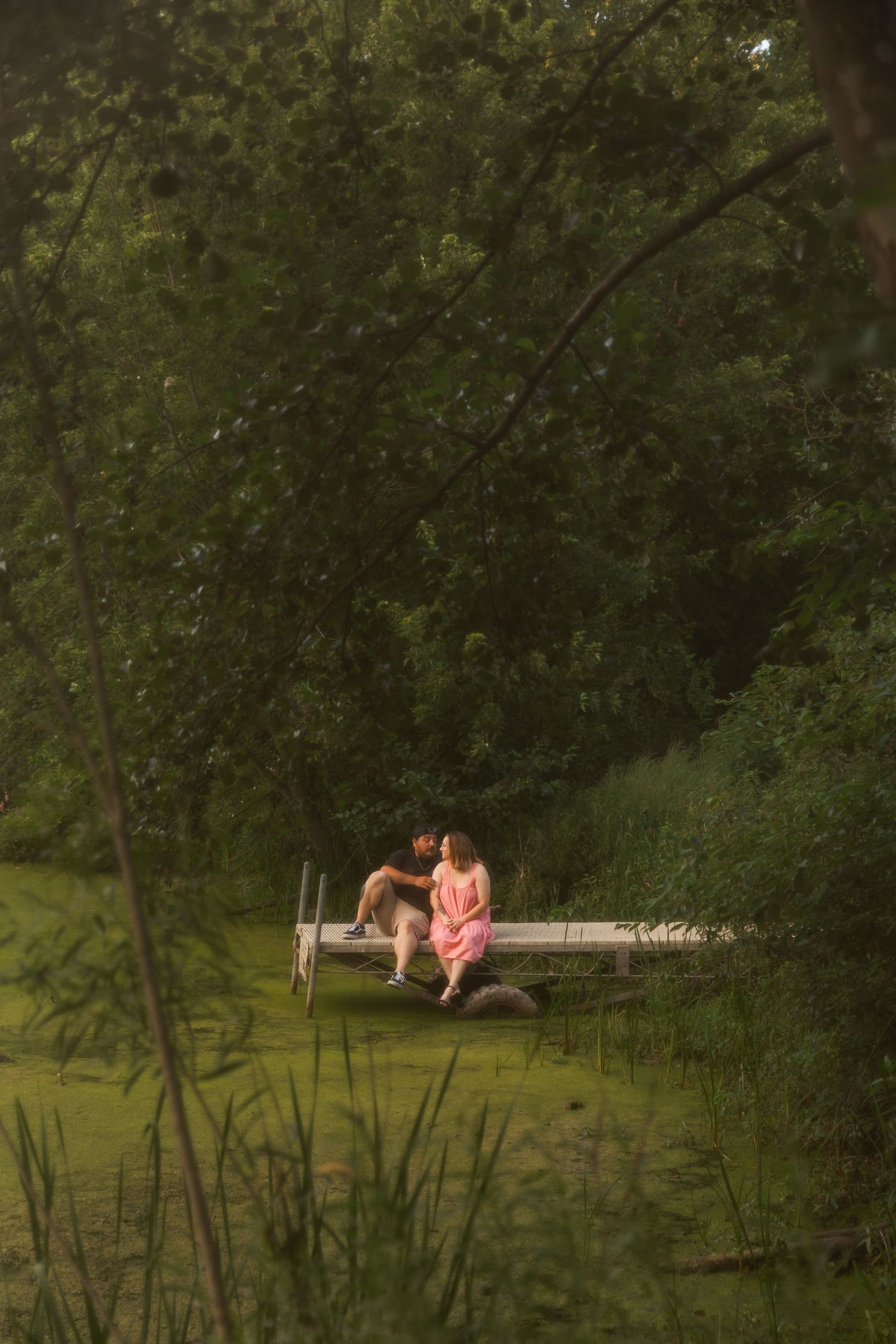 Romantic couple sitting together on a wooden dock surrounded by lush green trees reflected in calm water.