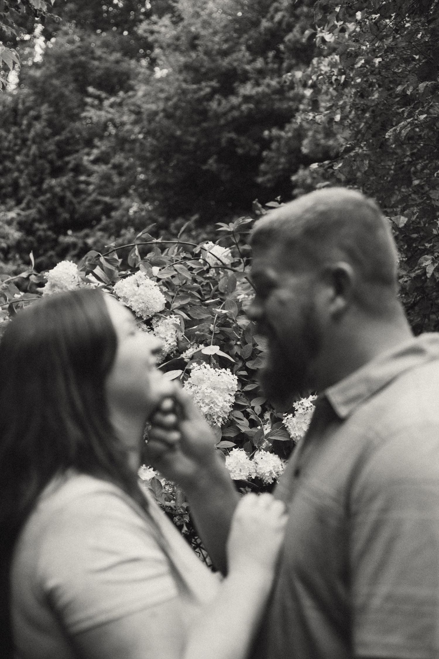 A black and white photo of a couple about to kiss near blooming white hydrangea flowers in a garden.
