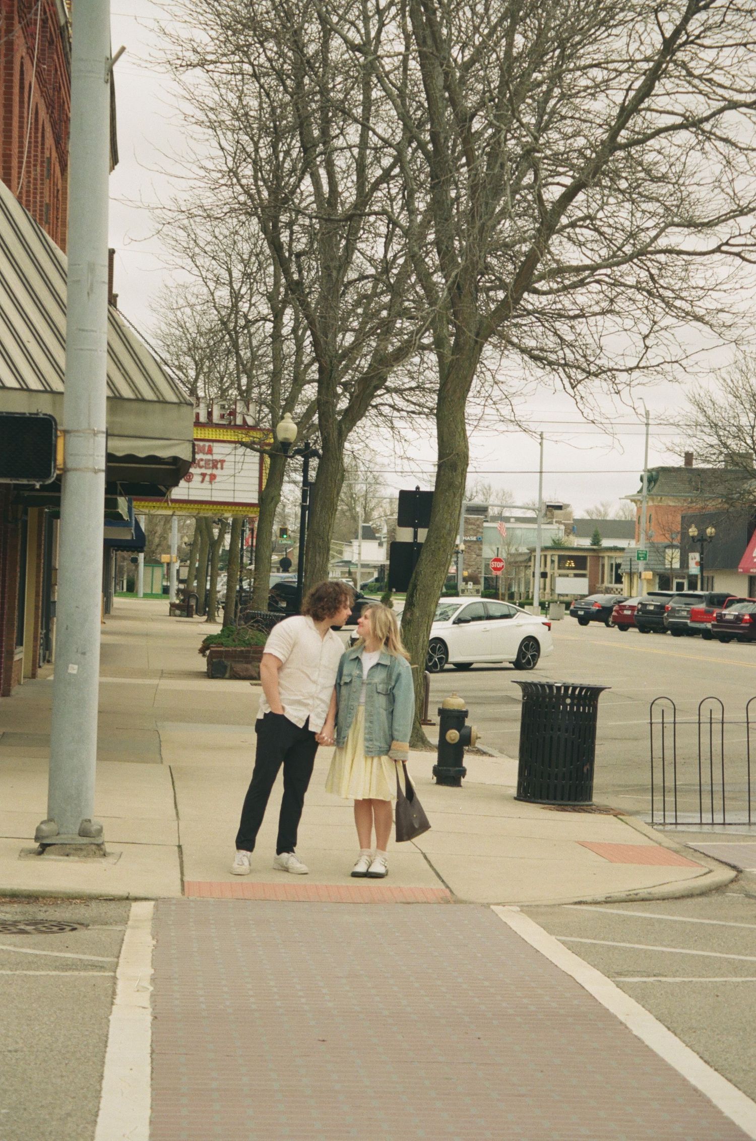 Couple holds hands on a quiet small-town sidewalk lined with bare trees and brick storefronts.