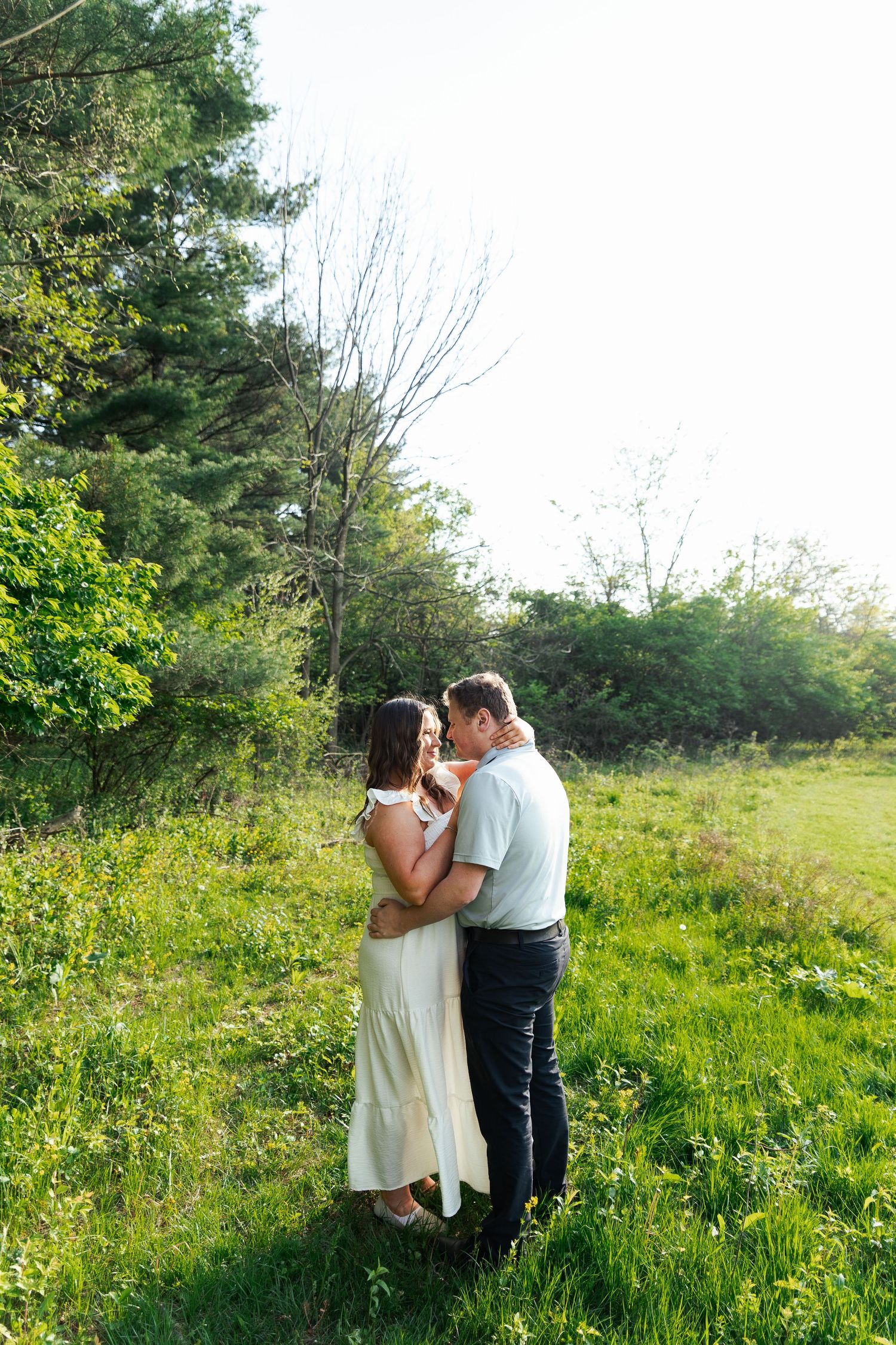 A couple embraces tenderly in a lush green meadow surrounded by trees in soft sunlight.