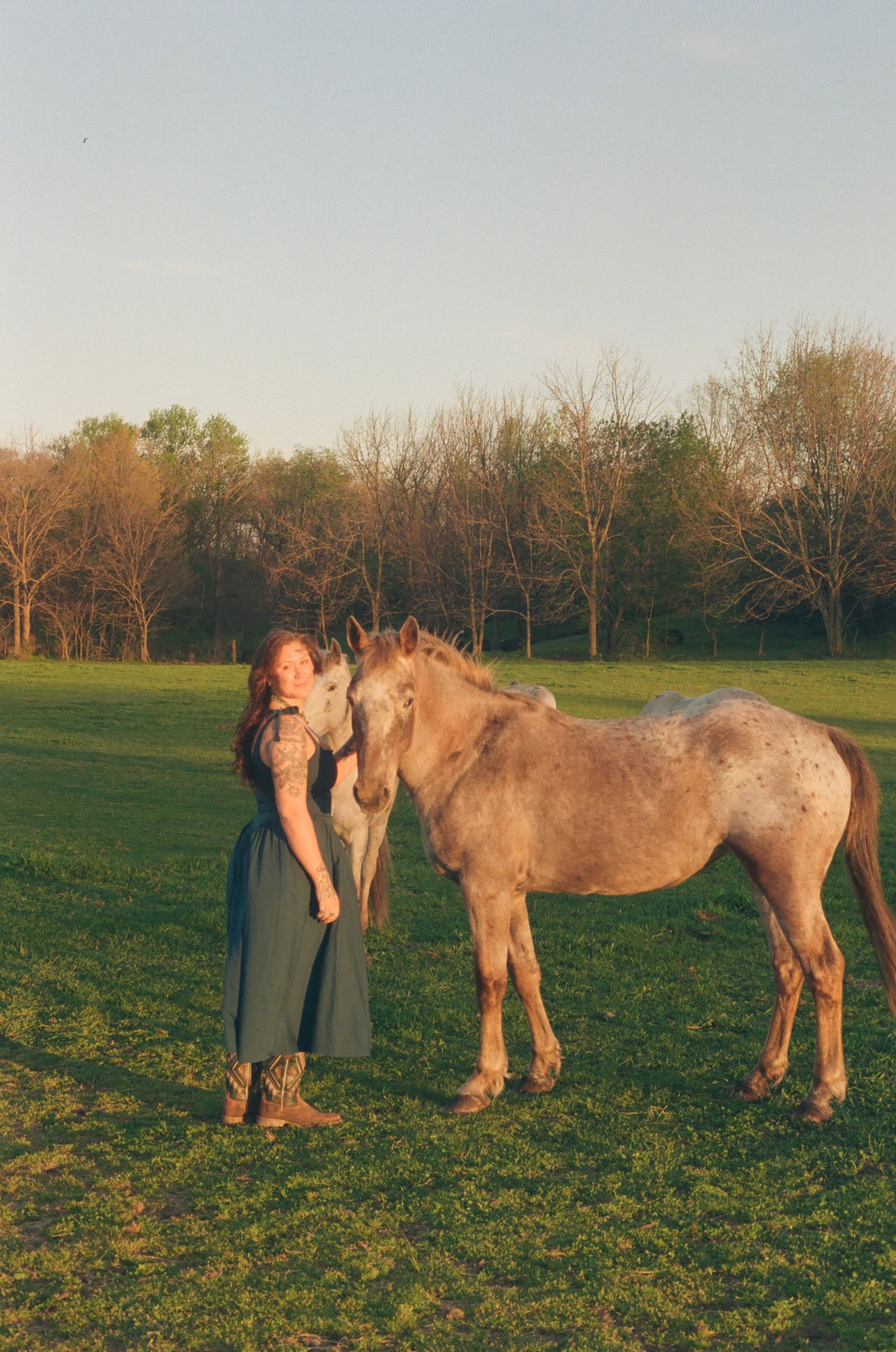 Young woman in teal dress kissing an Appaloosa horse in a green field at golden hour.
