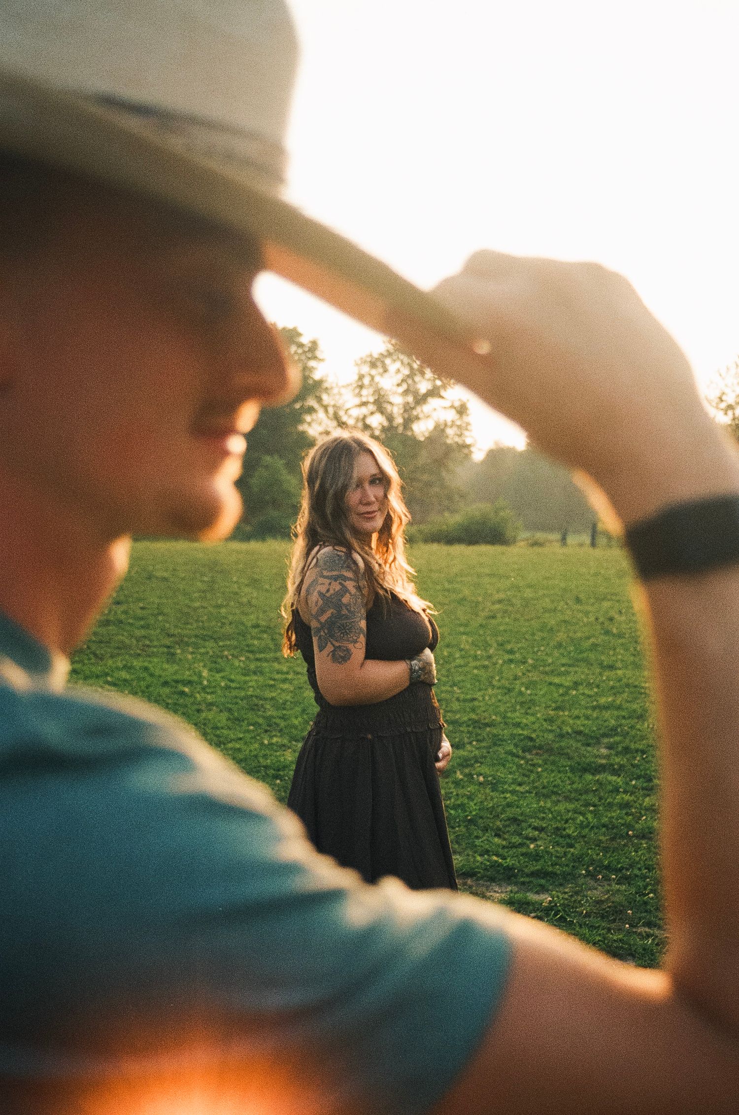A man in a hat tips his brim toward a tattooed woman in a black dress standing in a sunlit green park.