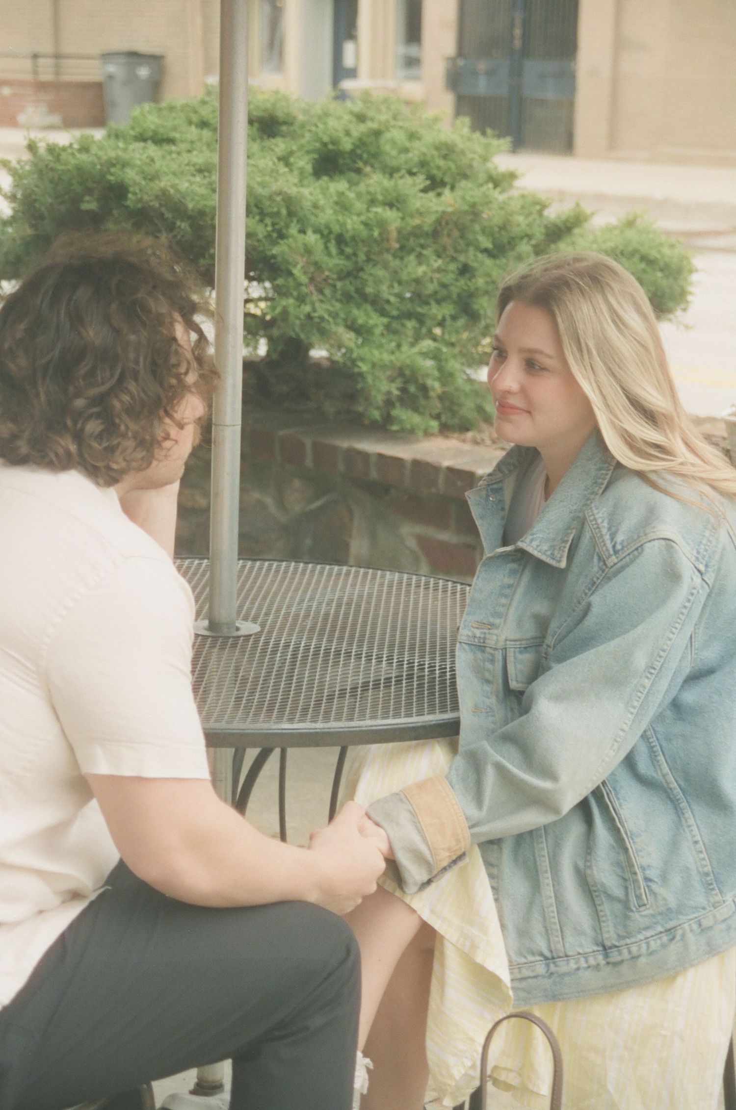 Young couple holding hands and talking intimately at outdoor cafe table with green shrubs behind.