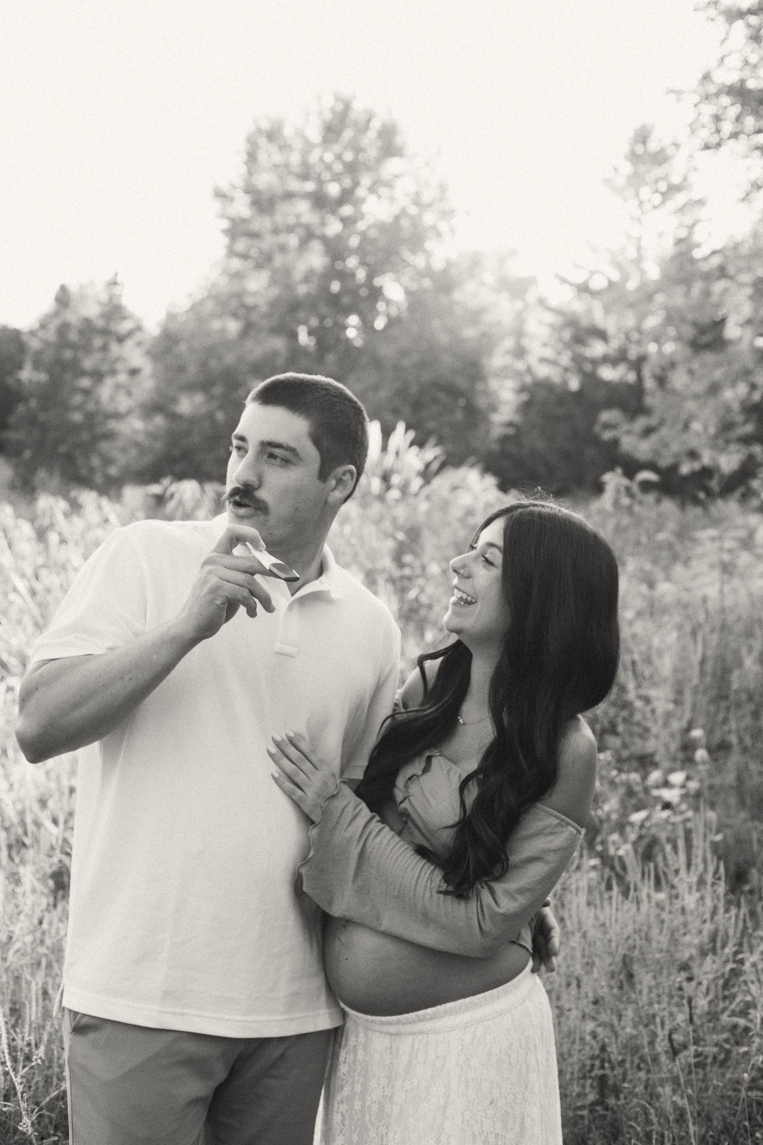 Black and white maternity photo of couple standing in wildflower field, woman smiling joyfully.