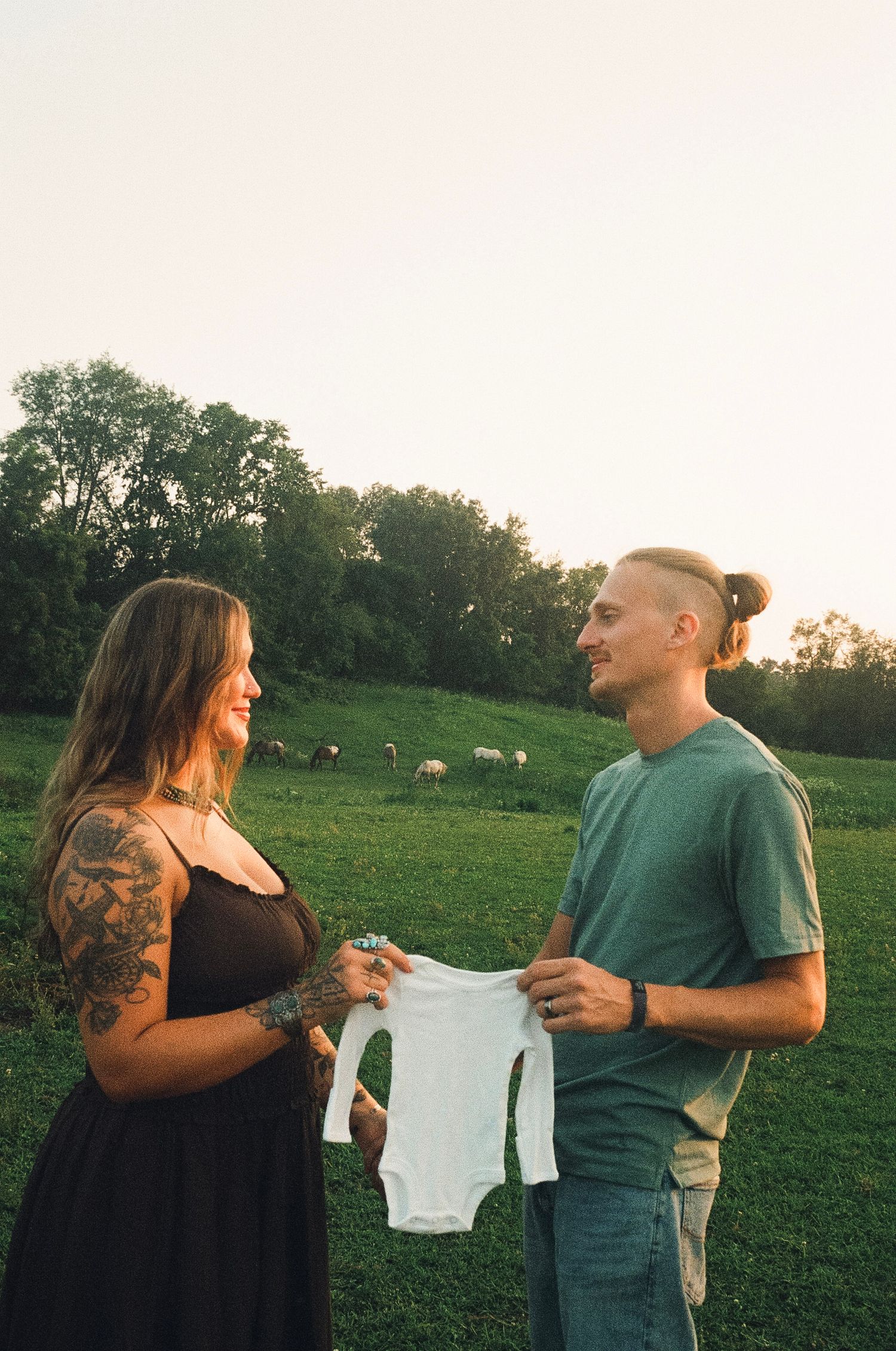 Couple holding white baby onesie in green field at sunset, pregnancy announcement photo with tattoos and casual attire.