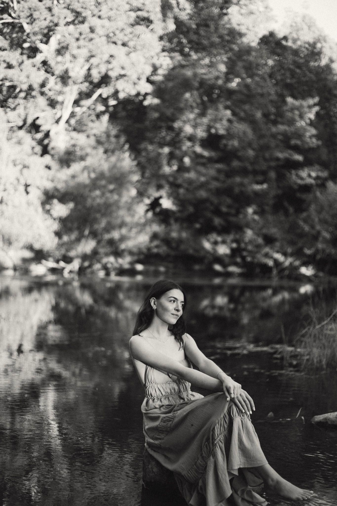 Black and white portrait of serene woman sitting barefoot by a tranquil forest pond.