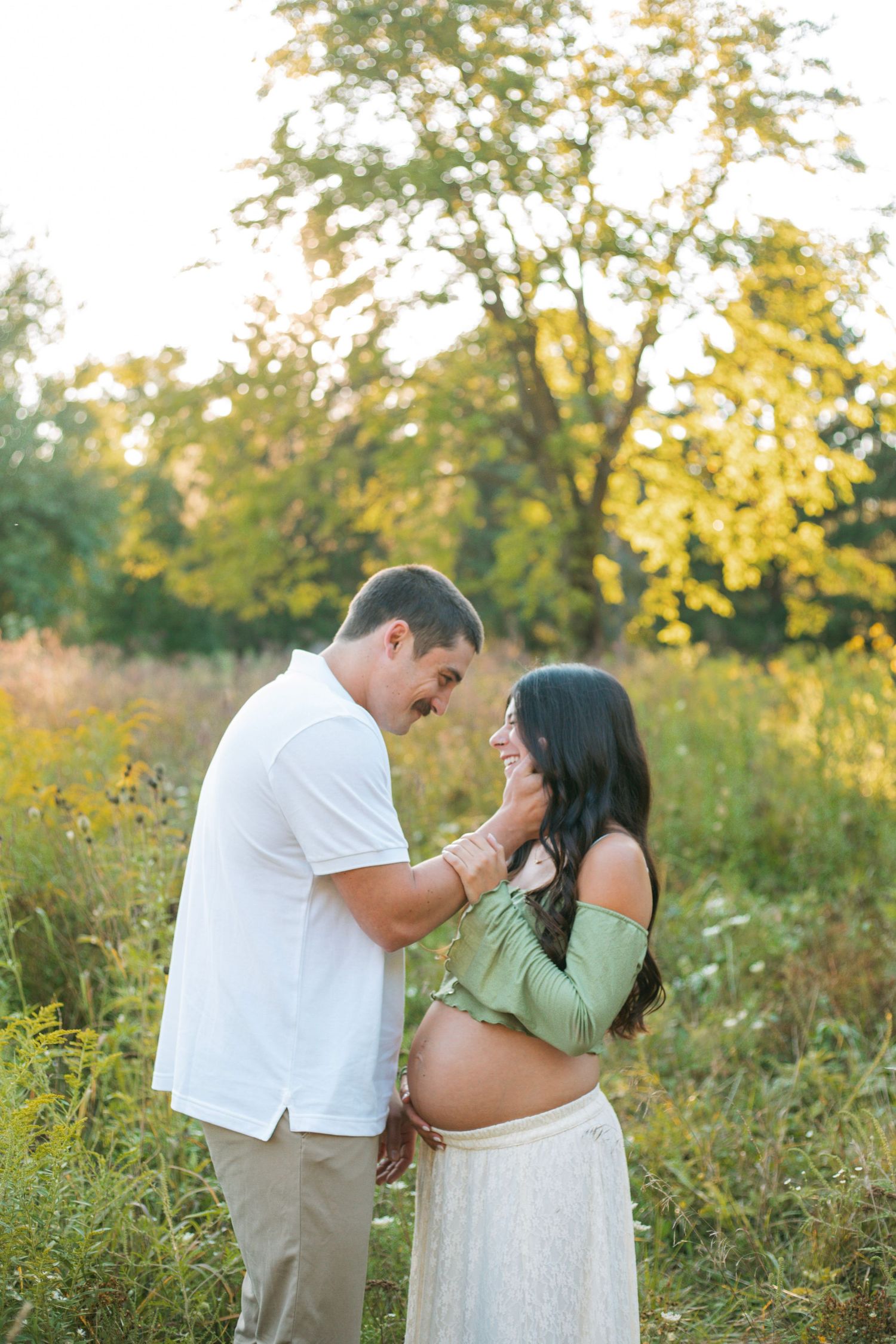 Couple sharing tender moment in sunlit meadow, pregnant woman in green crop top and white lace skirt.