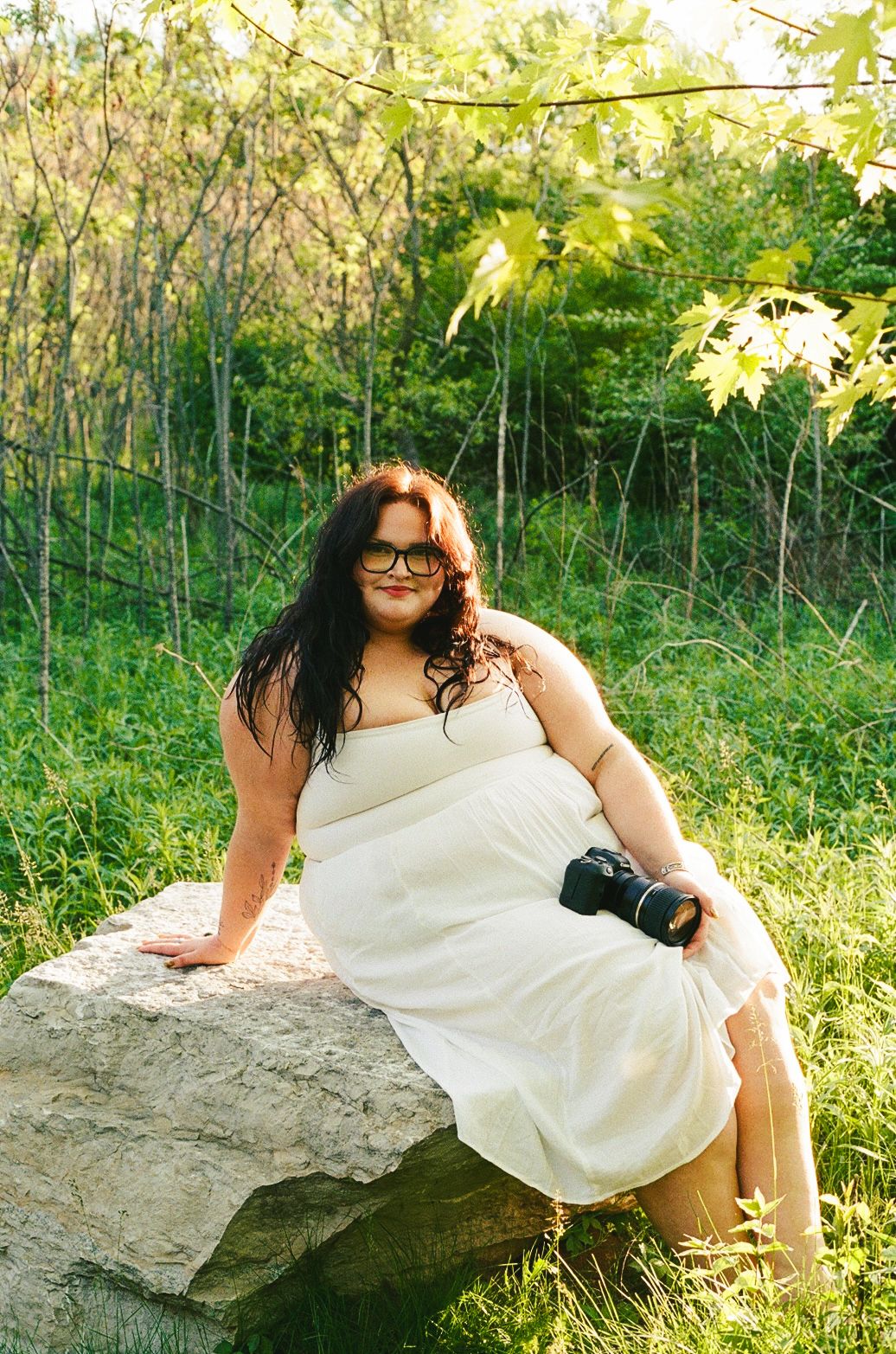 Woman in white dress sitting on a rock holding a camera in a lush green setting.