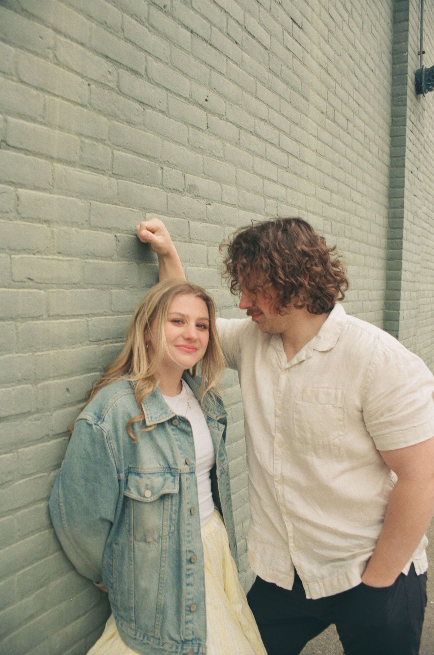 Couple leaning against a green brick wall smiling, woman in denim jacket, man in linen shirt.