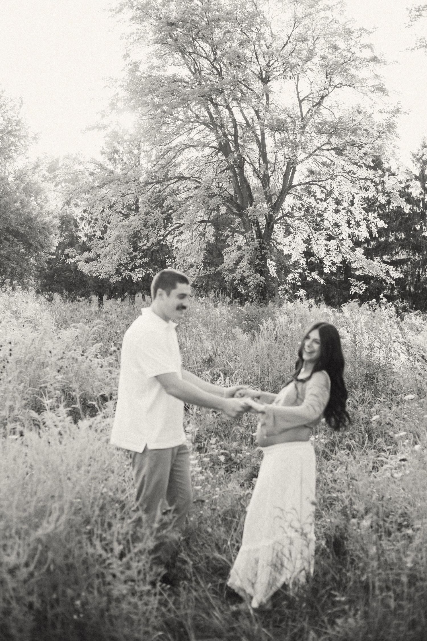Black and white maternity photo of couple holding hands and dancing in a wildflower meadow.