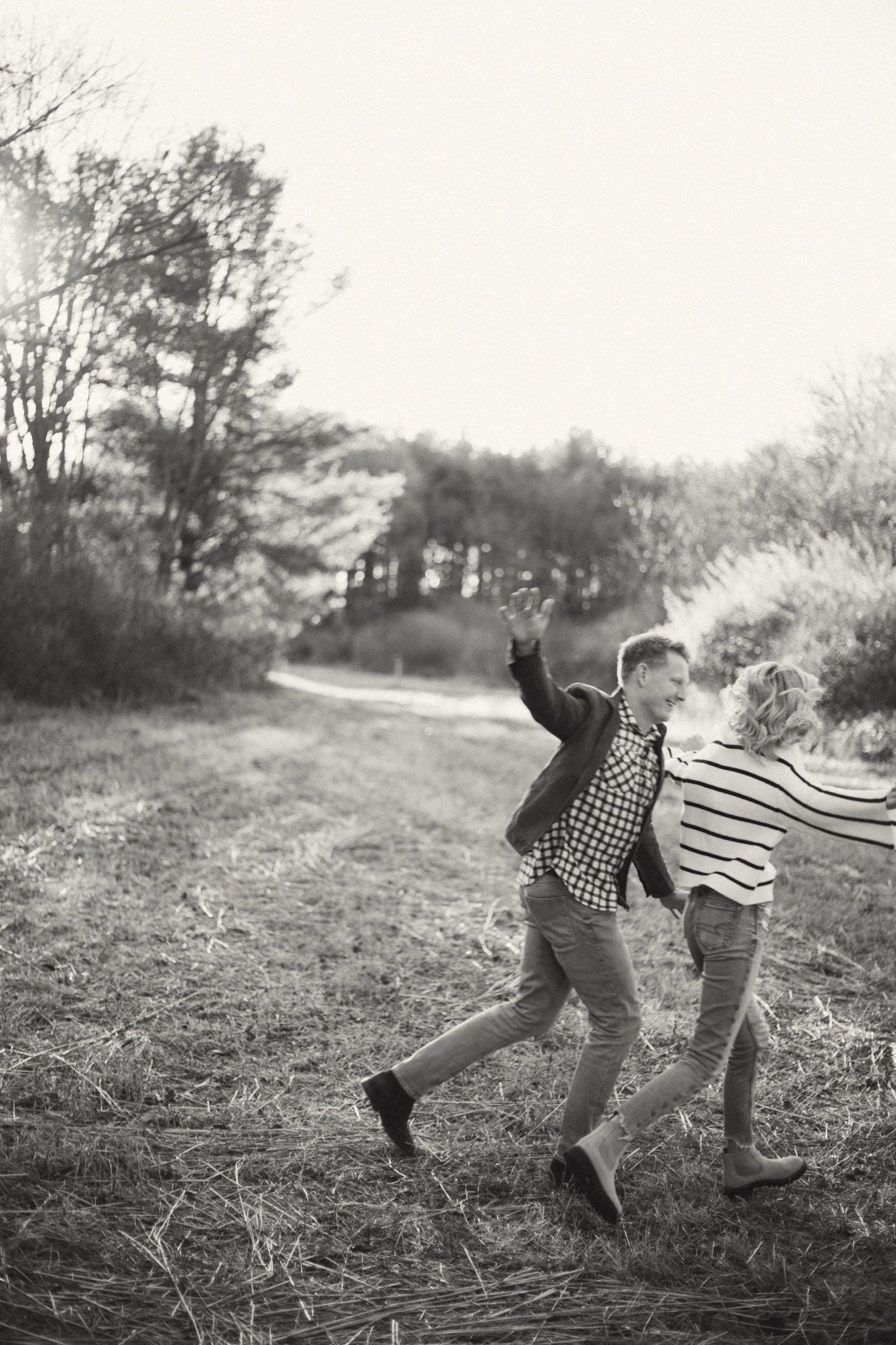Black and white photo of playful couple running through autumn field along scenic wooded path.
