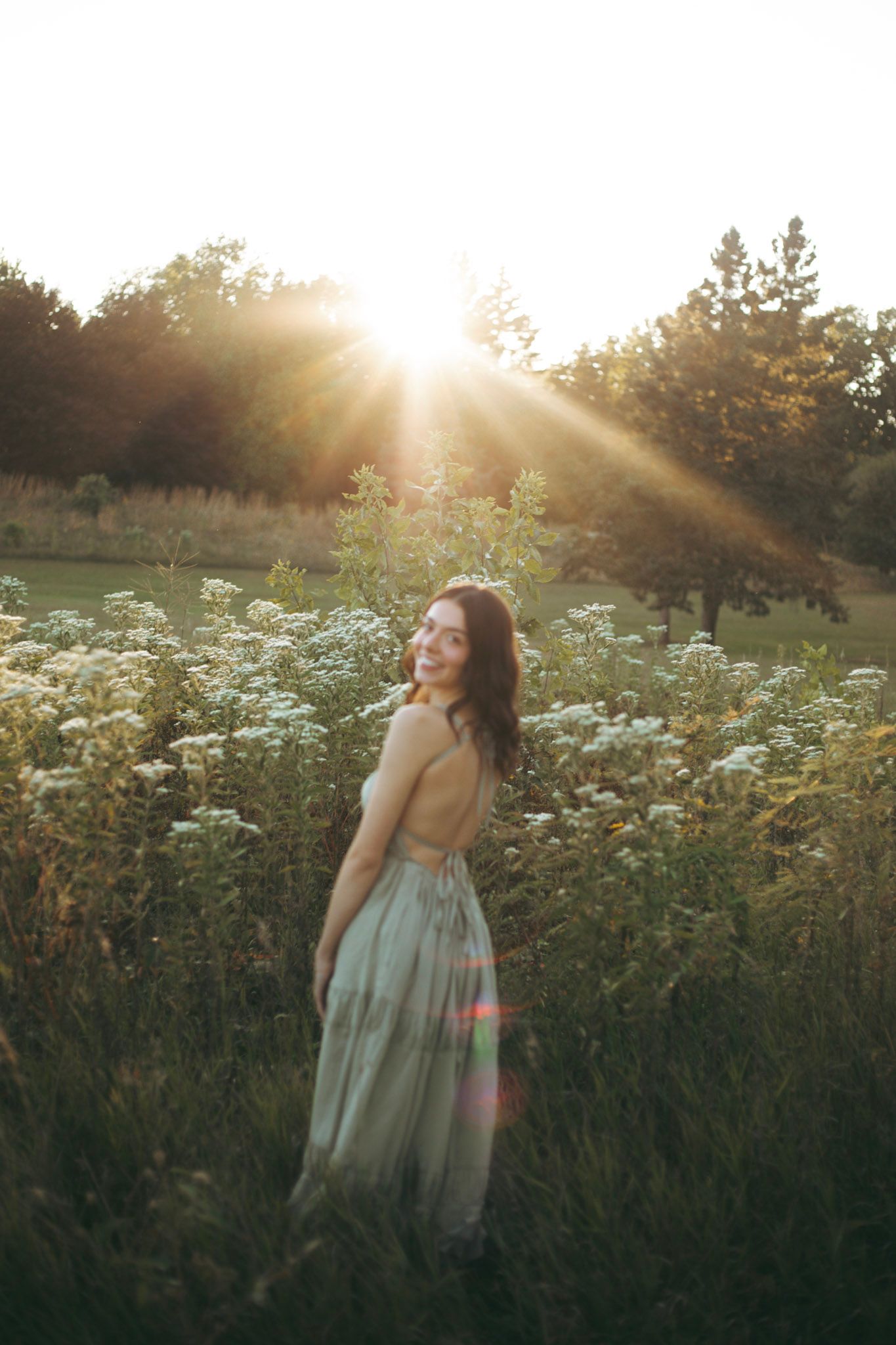 Young woman in backless sage dress standing in wildflower field bathed in golden sunset rays.
