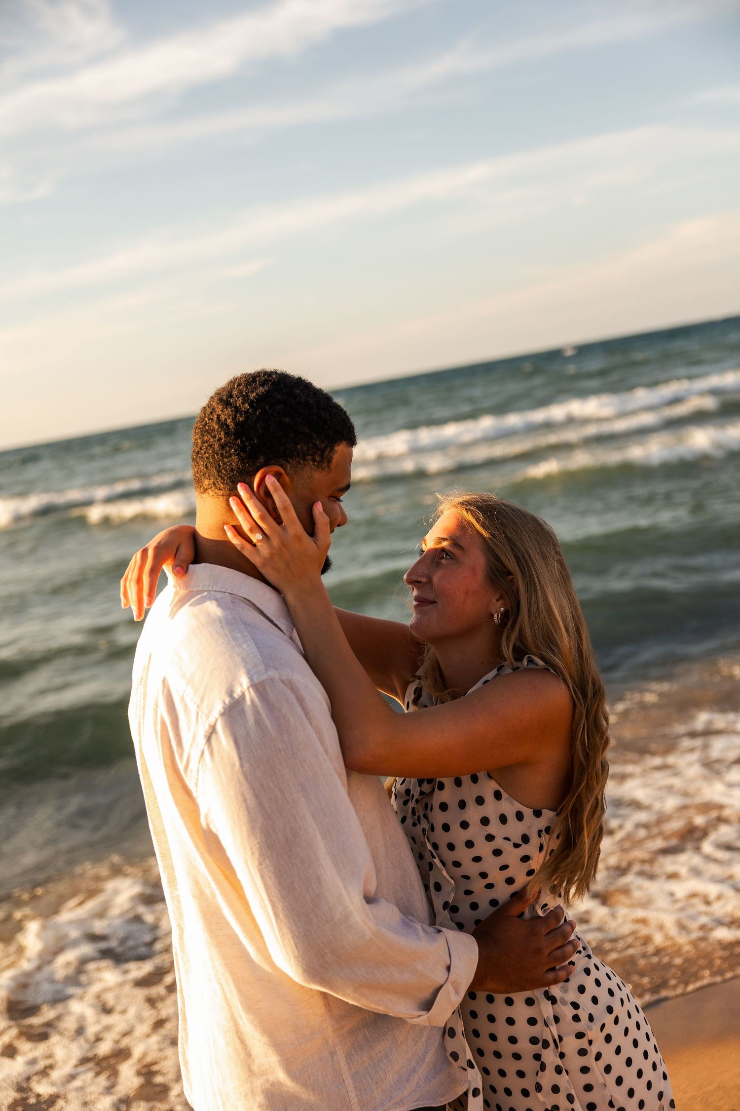 A romantic couple embraces on a sunny beach at sunset, with ocean waves in the background.