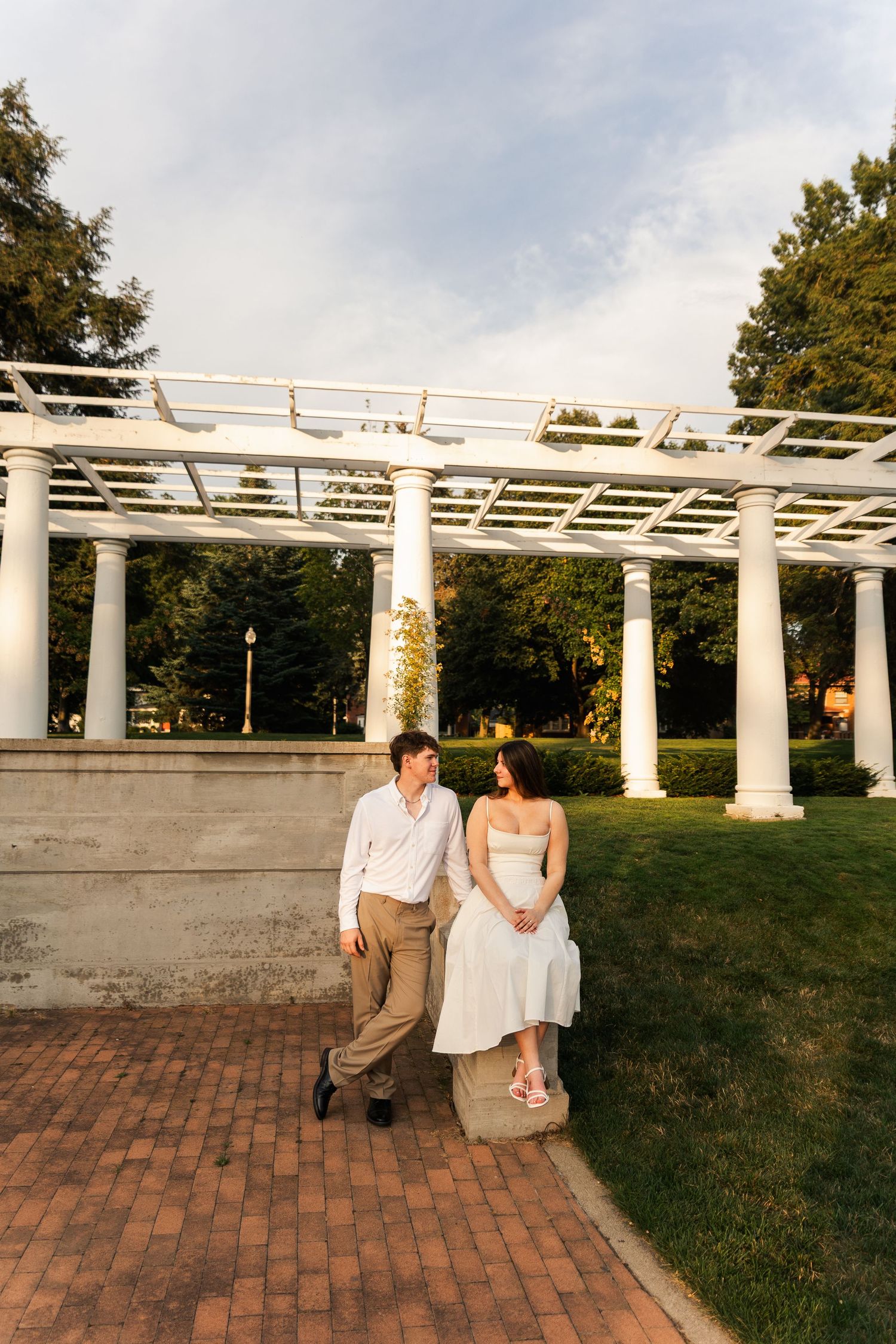 Engaged couple relaxing together near white pergola columns in a manicured park at golden hour sunset.