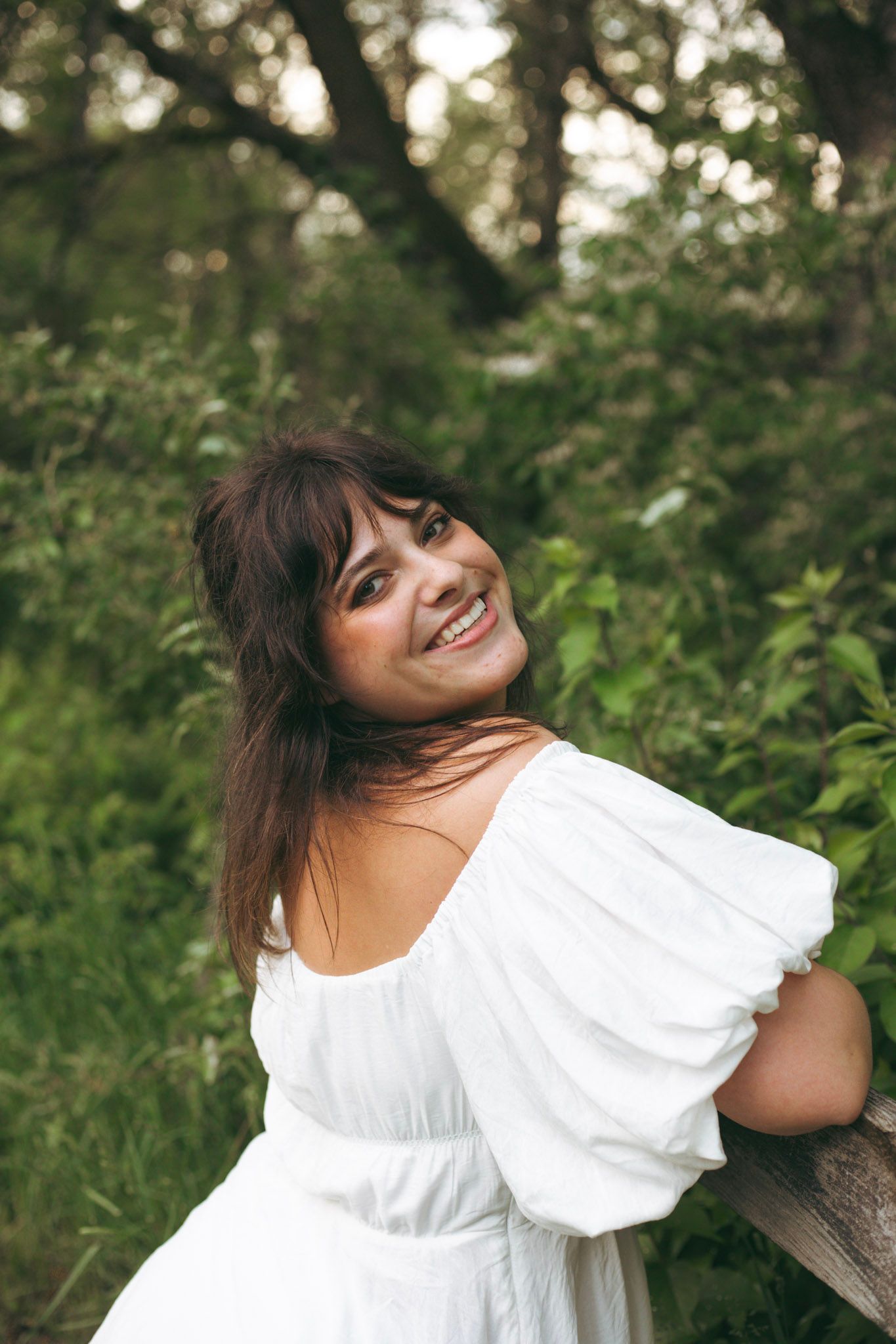 Cheerful brunette woman in white off-shoulder dress smiling over shoulder among lush green foliage.