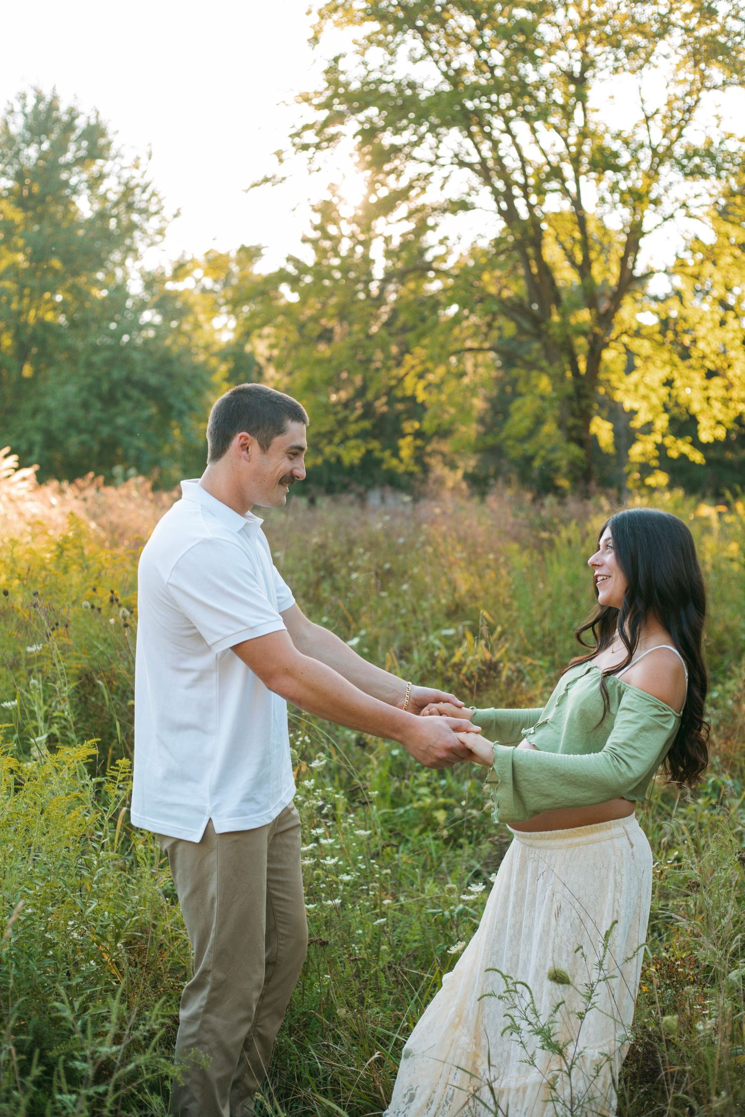 Smiling expectant couple holding hands facing each other in a lush green field at golden hour.