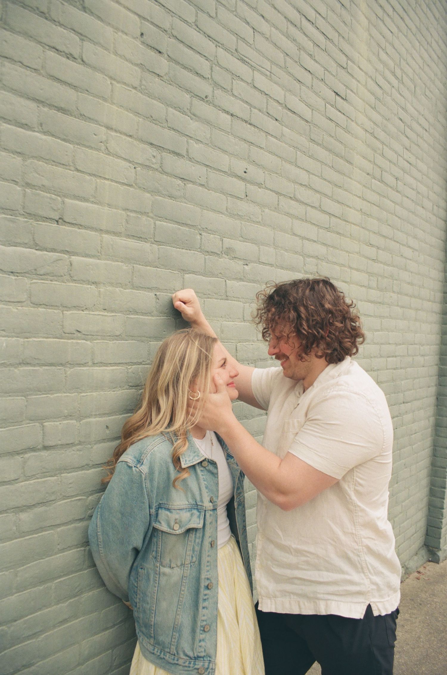 Couple sharing a playful moment against a green brick wall, man gently touching womans face.