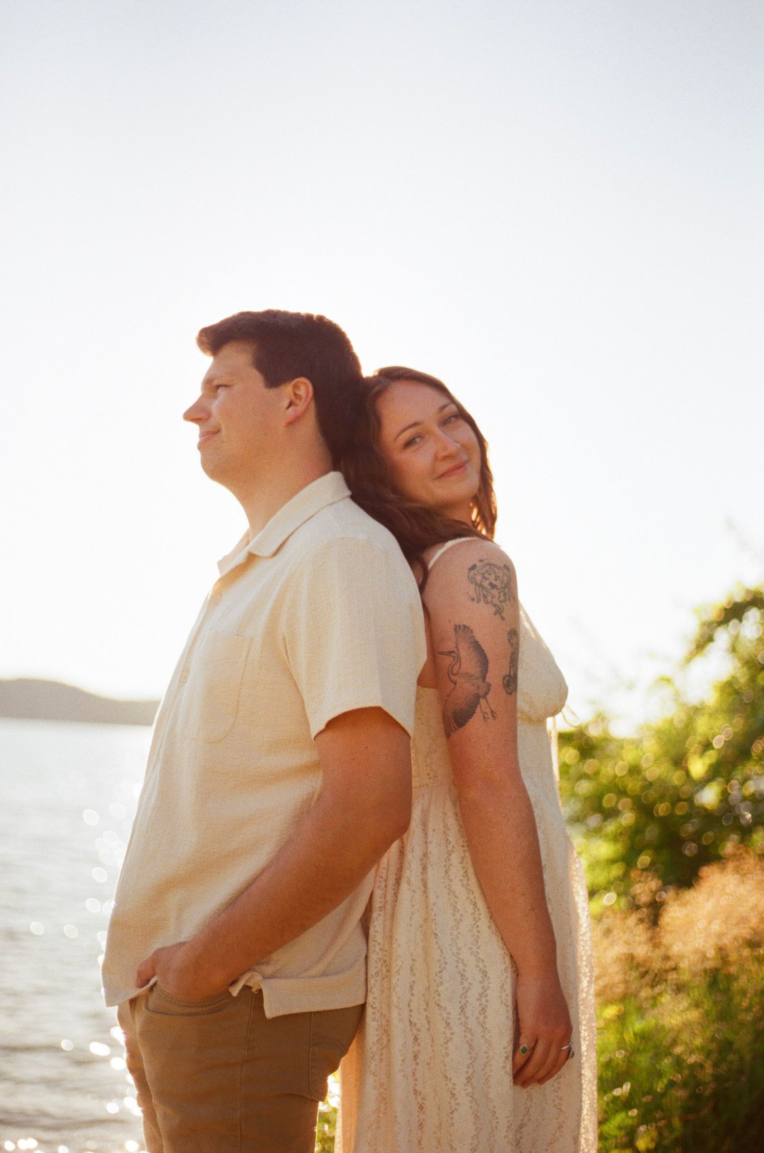 Couple standing back-to-back at golden hour near water, woman with tattoos in white lace dress smiling warmly.