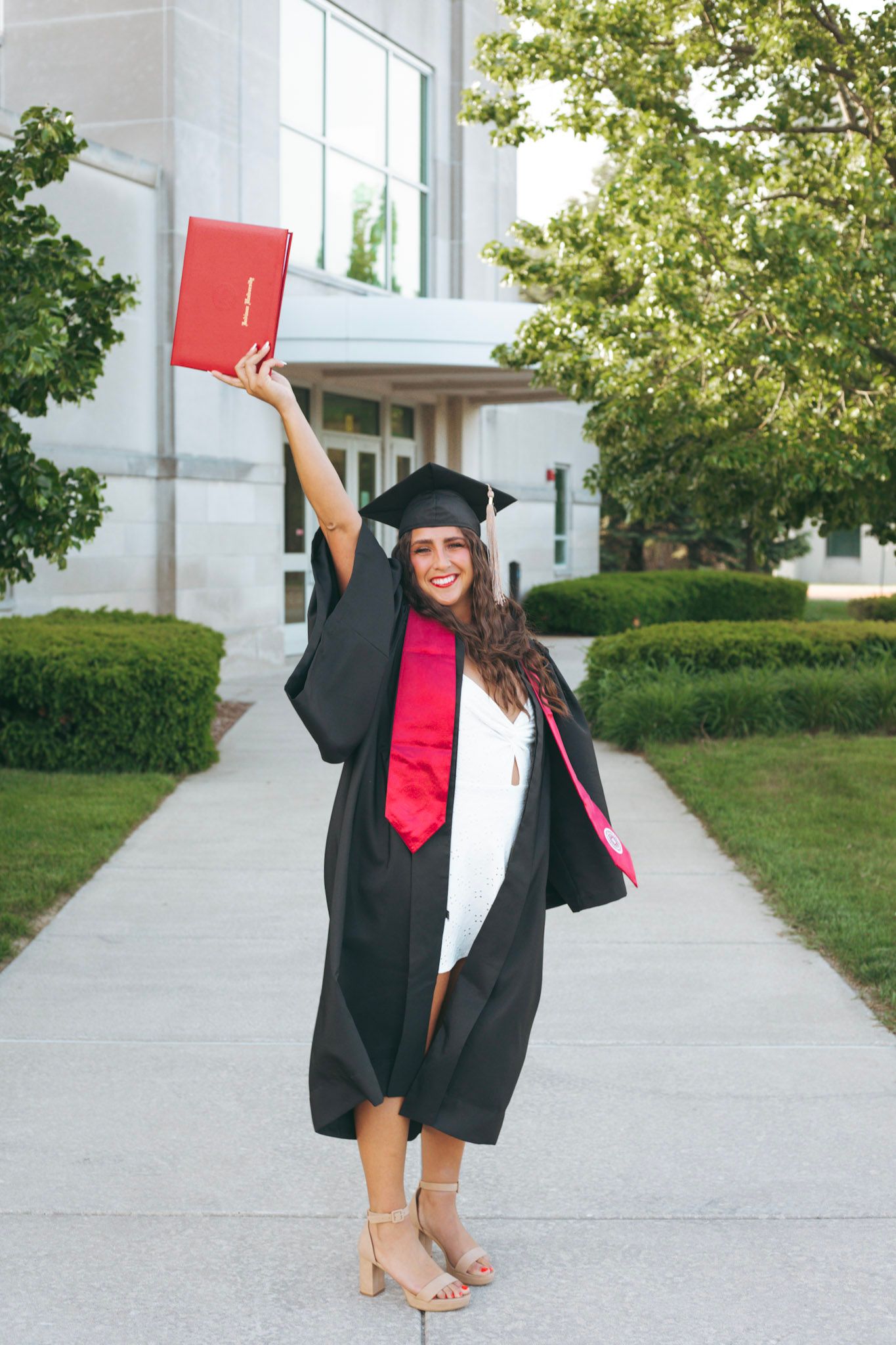 Excited female graduate in black cap and gown raising red diploma in the air outside campus building.