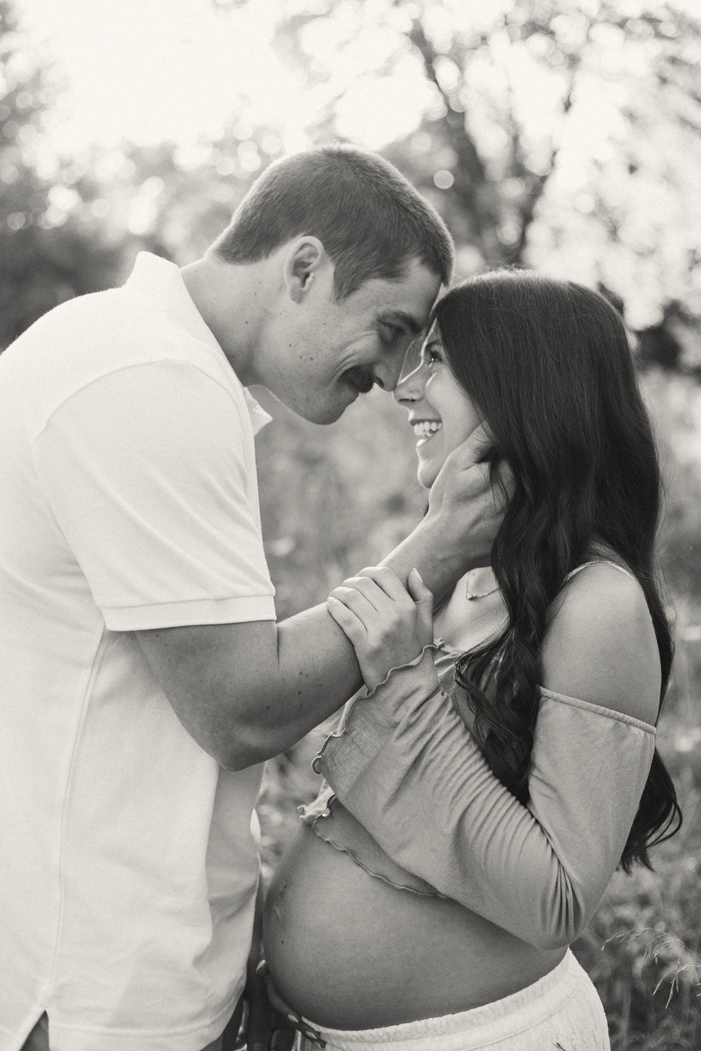 Black and white photo of smiling pregnant couple touching foreheads tenderly outdoors.