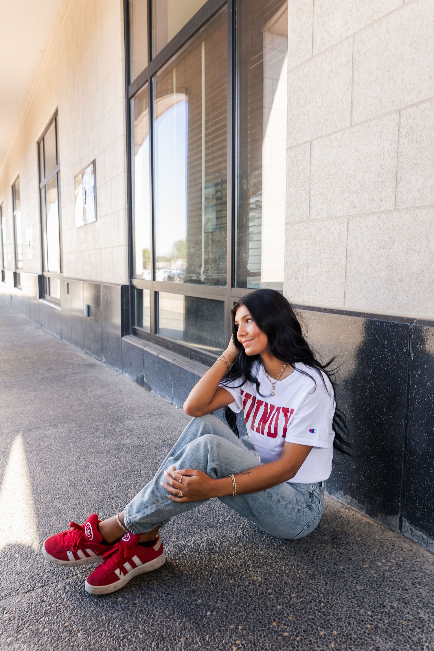 Young woman in white Champion tee and jeans sitting against a building wall in red sneakers.