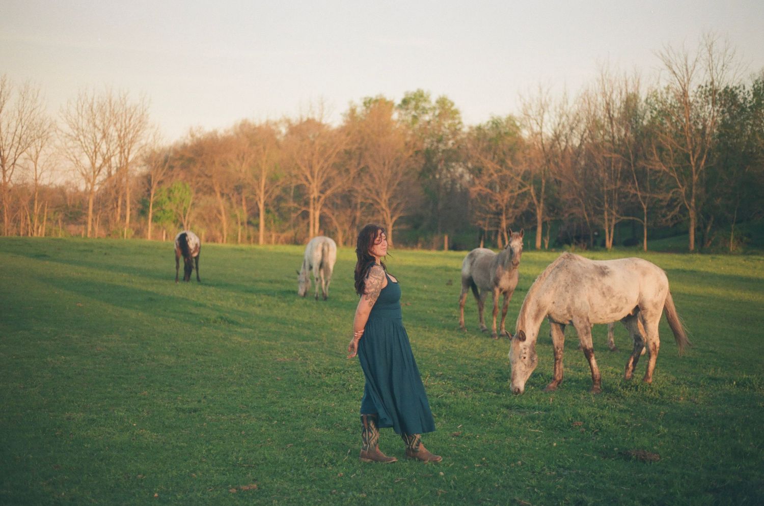Woman in teal dress stands serenely among white horses grazing in a sunlit green meadow.