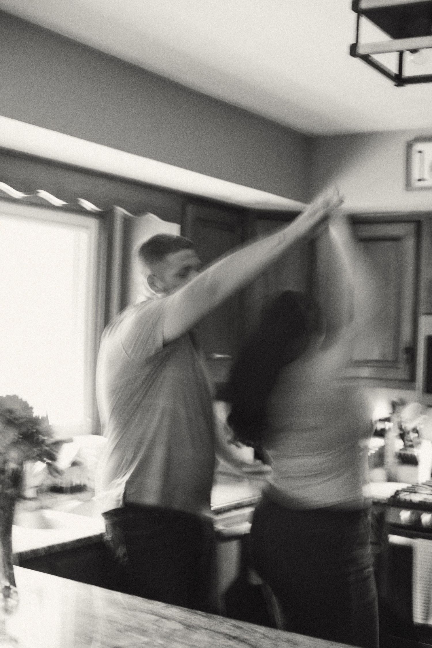 Blurred black and white photo of a couple dancing joyfully together in a cozy home kitchen.