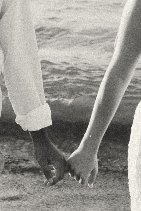 Black and white photo of an interracial couple holding hands on a sandy beach near the ocean.