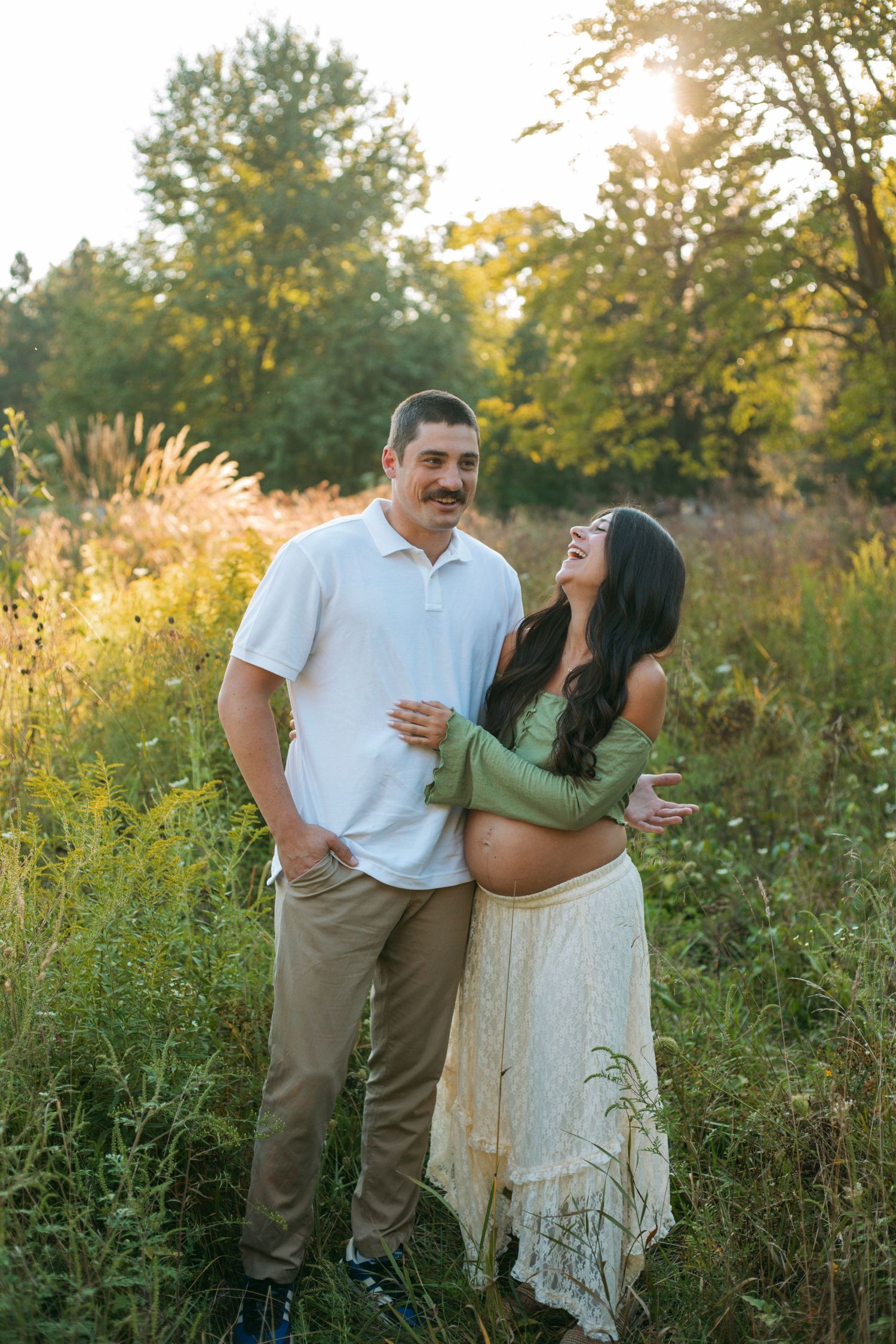 Happy couple laughing together in golden hour meadow, pregnant woman in green top and white skirt.