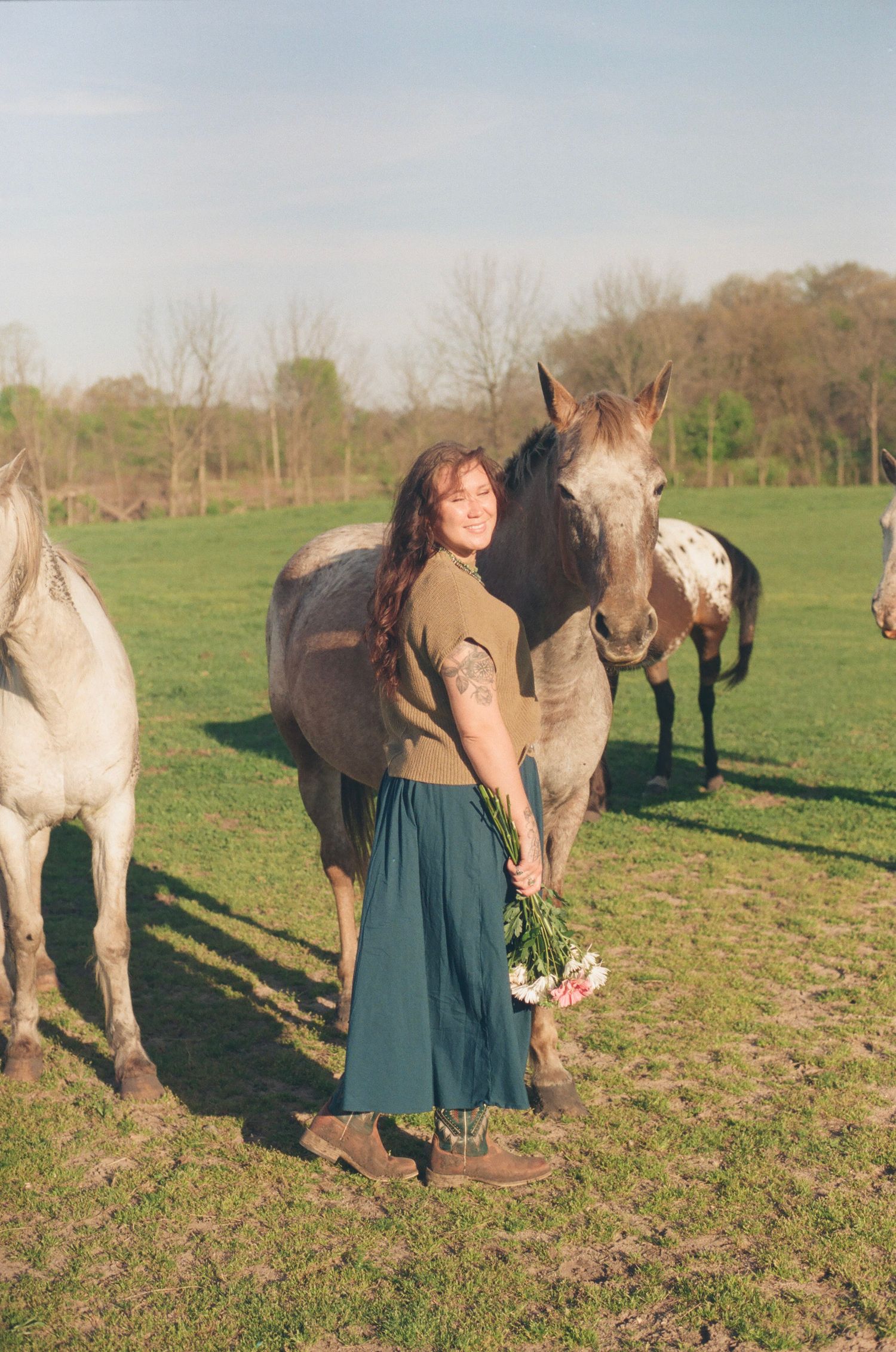 Smiling woman holds pink flowers beside a dappled horse in a sunny green pasture.