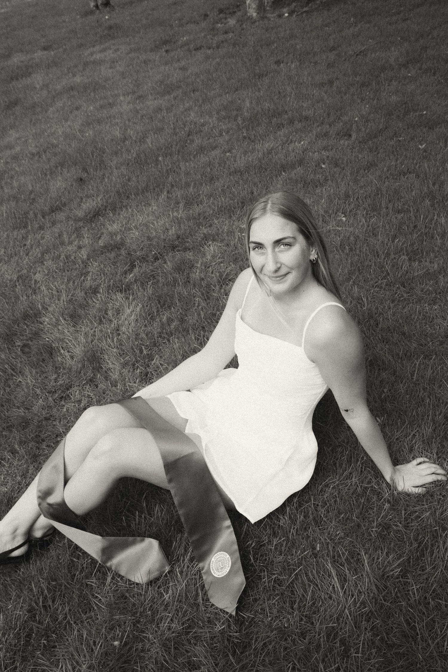 Black and white photo of a smiling young woman in a white dress sitting on a grassy hill.