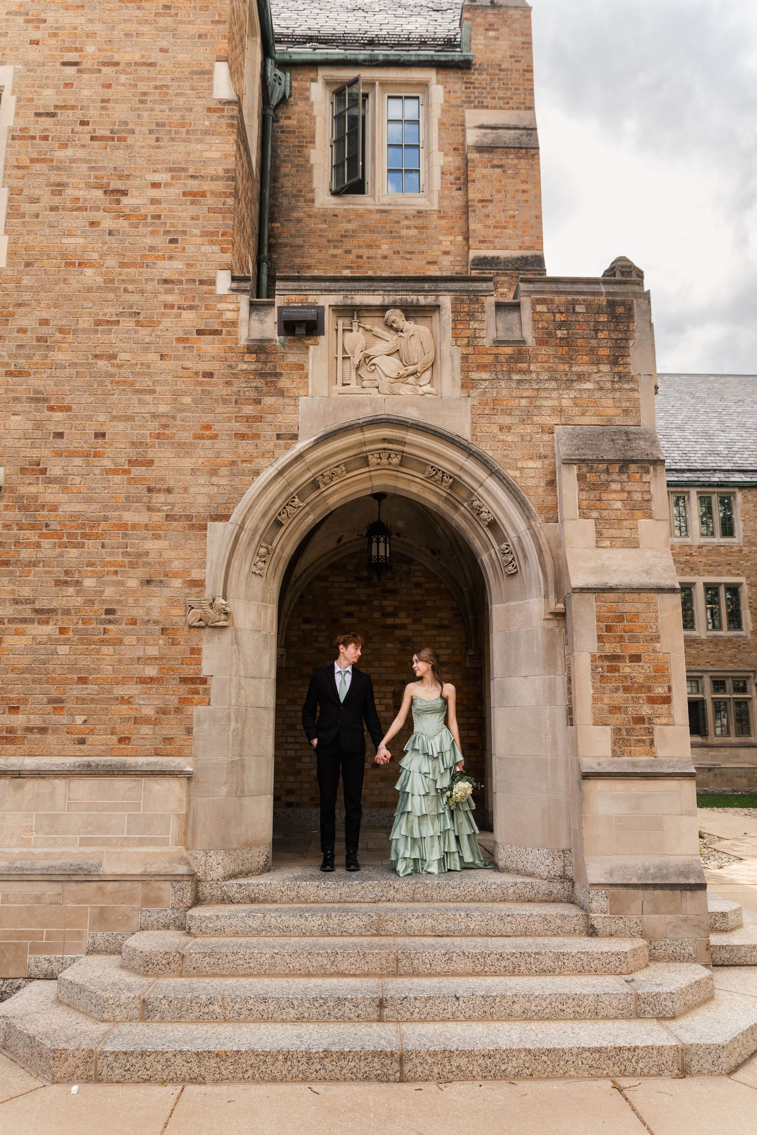 Elegant couple holding hands under a stone archway of a historic brick building.