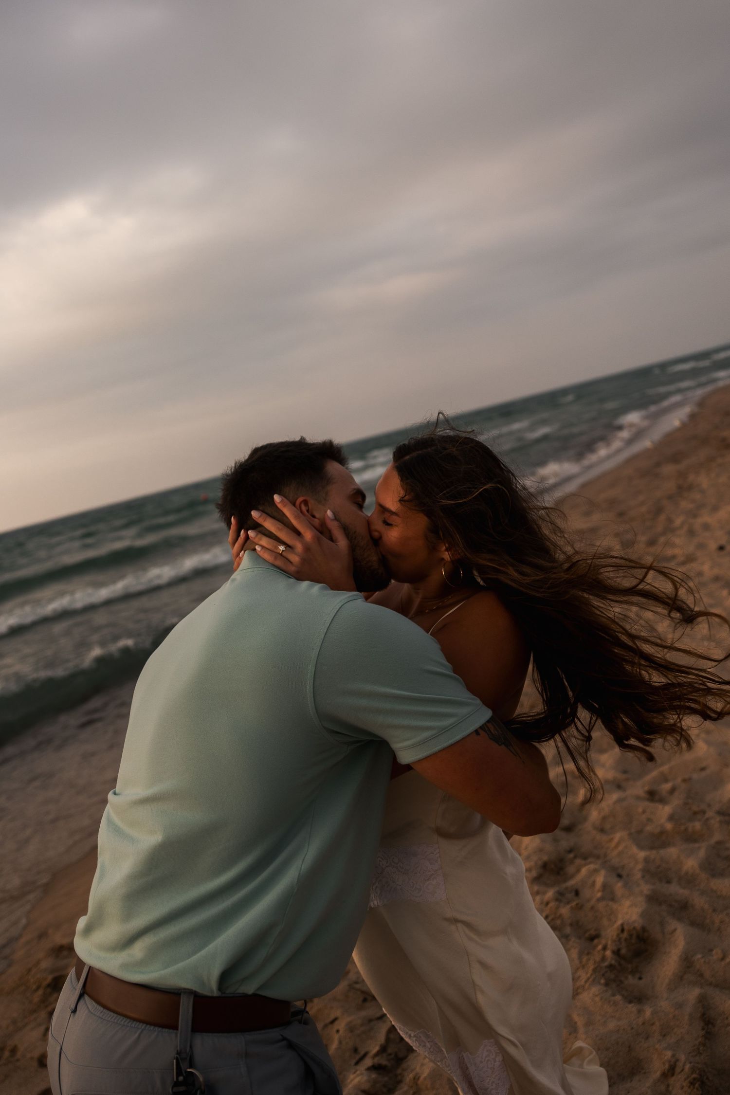 Couple sharing a romantic kiss on a sandy beach at sunset with ocean waves and cloudy sky in the background.