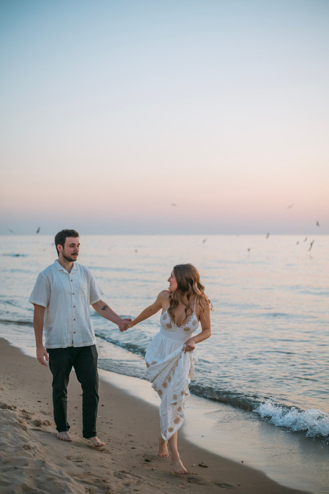 Couple holding hands and walking along a sandy beach at pastel sunset.