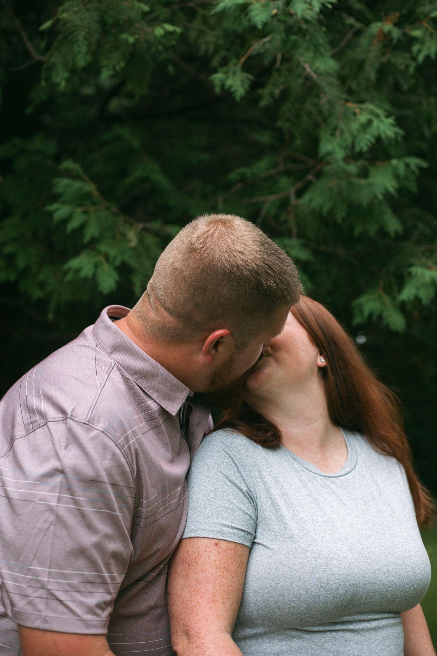 A couple shares a tender kiss outdoors with a lush green evergreen tree in the background.