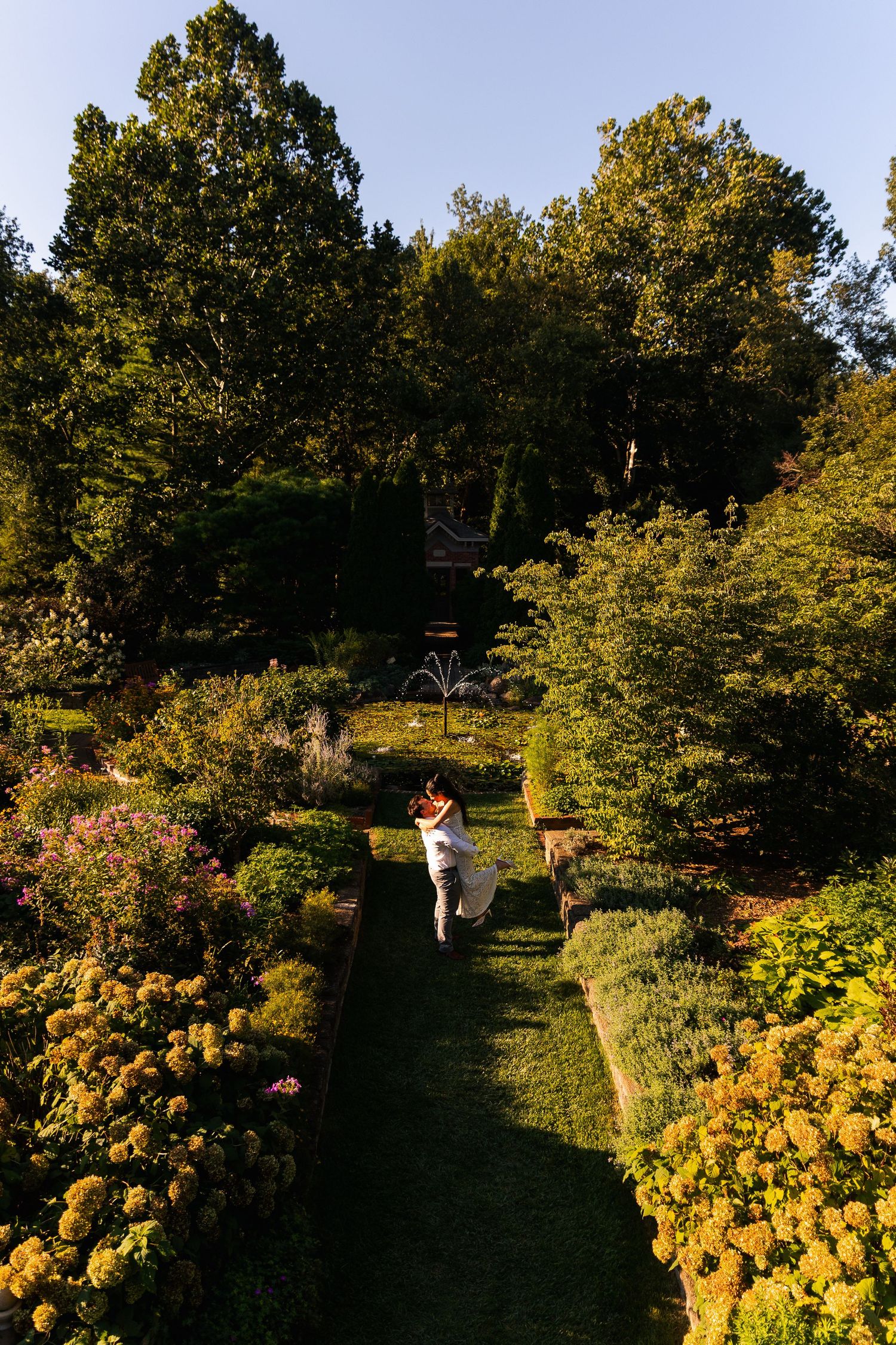 Couple embracing in a lush garden pathway surrounded by colorful yellow and pink flowers and tall green trees.