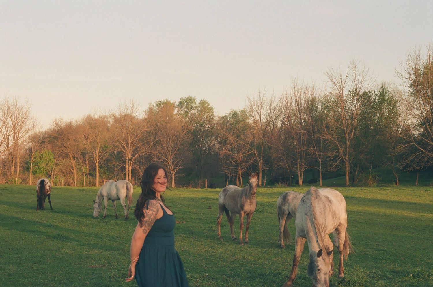 Woman in teal dress smiling among grazing horses in a golden hour farm pasture.