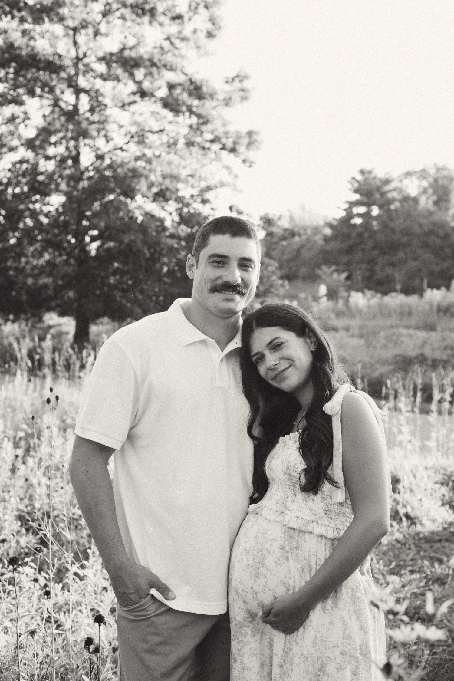 Black and white maternity photo of couple embracing in wildflower field, woman leaning on partners shoulder.