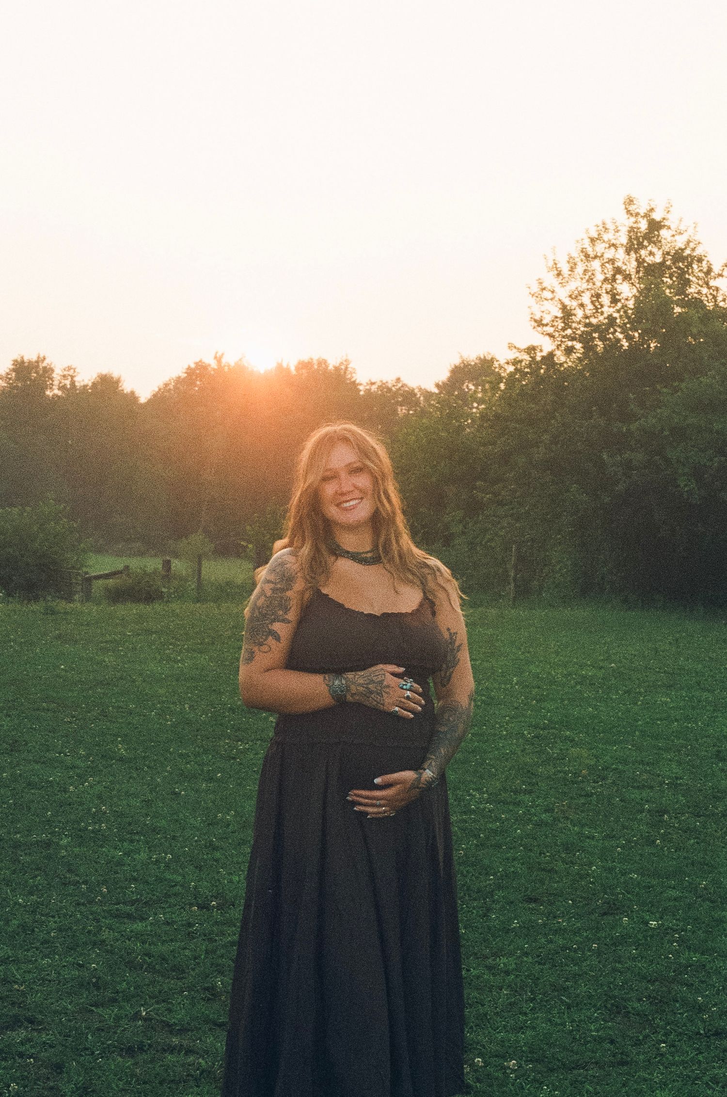 Smiling pregnant woman with tattoos in black maxi dress poses outdoors at golden hour sunset in green field.
