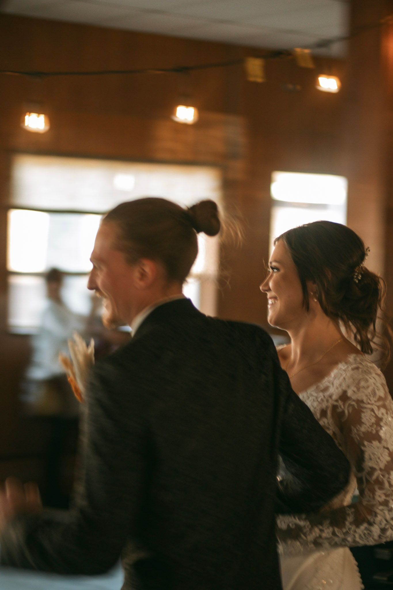 Smiling bride in lace gown beside groom with bun hairstyle at warmly lit reception.