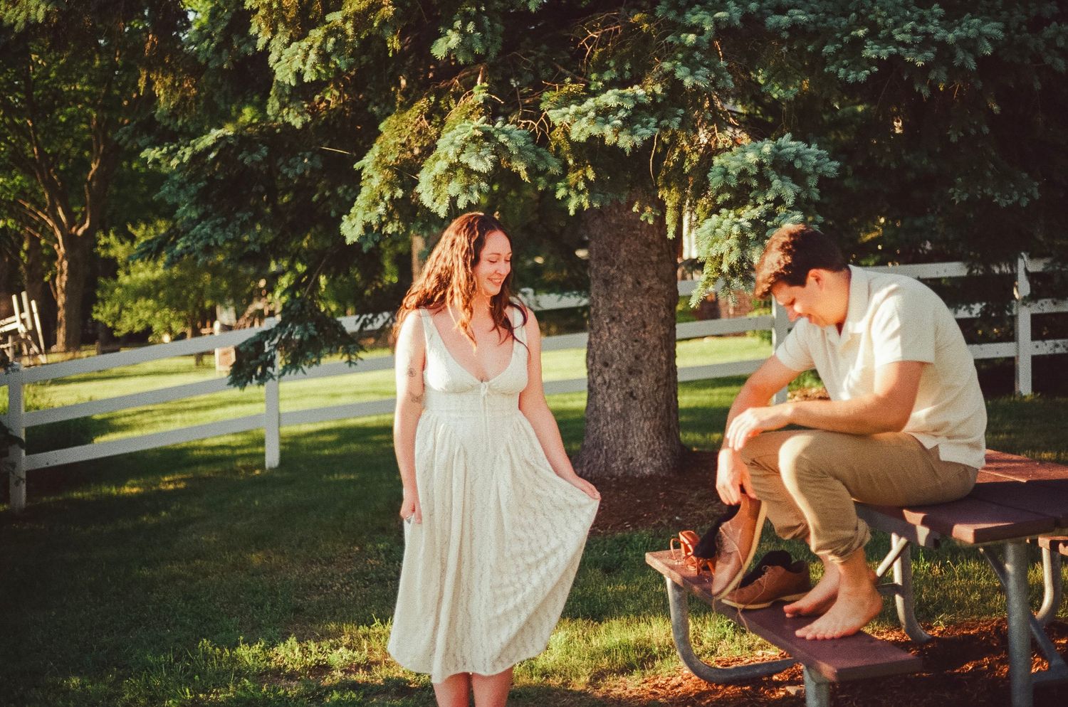 A woman in a white dress and a man sitting on a picnic table share a romantic moment in a sunny park.
