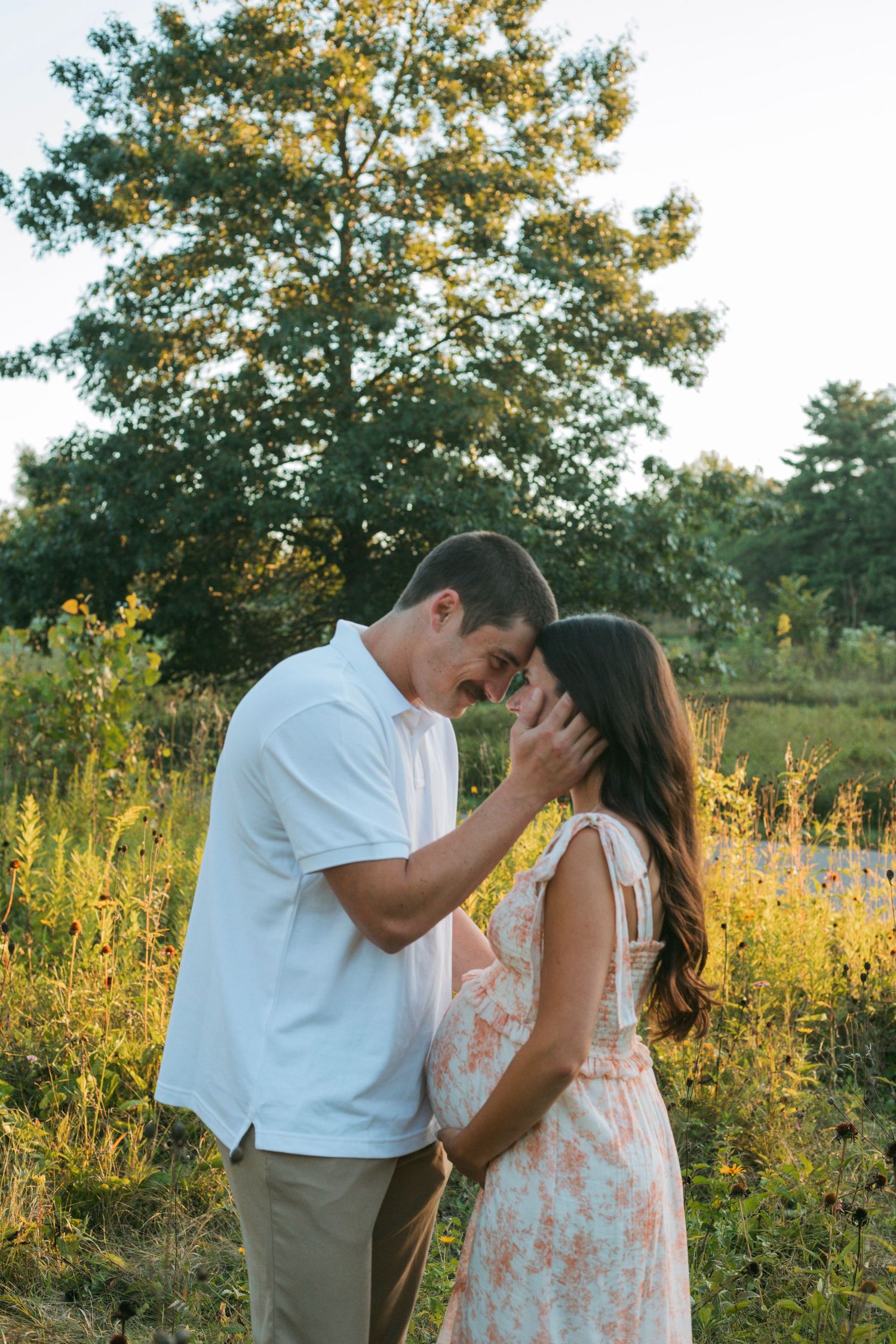 Couple sharing intimate forehead touch moment in golden hour wildflower field during maternity session.