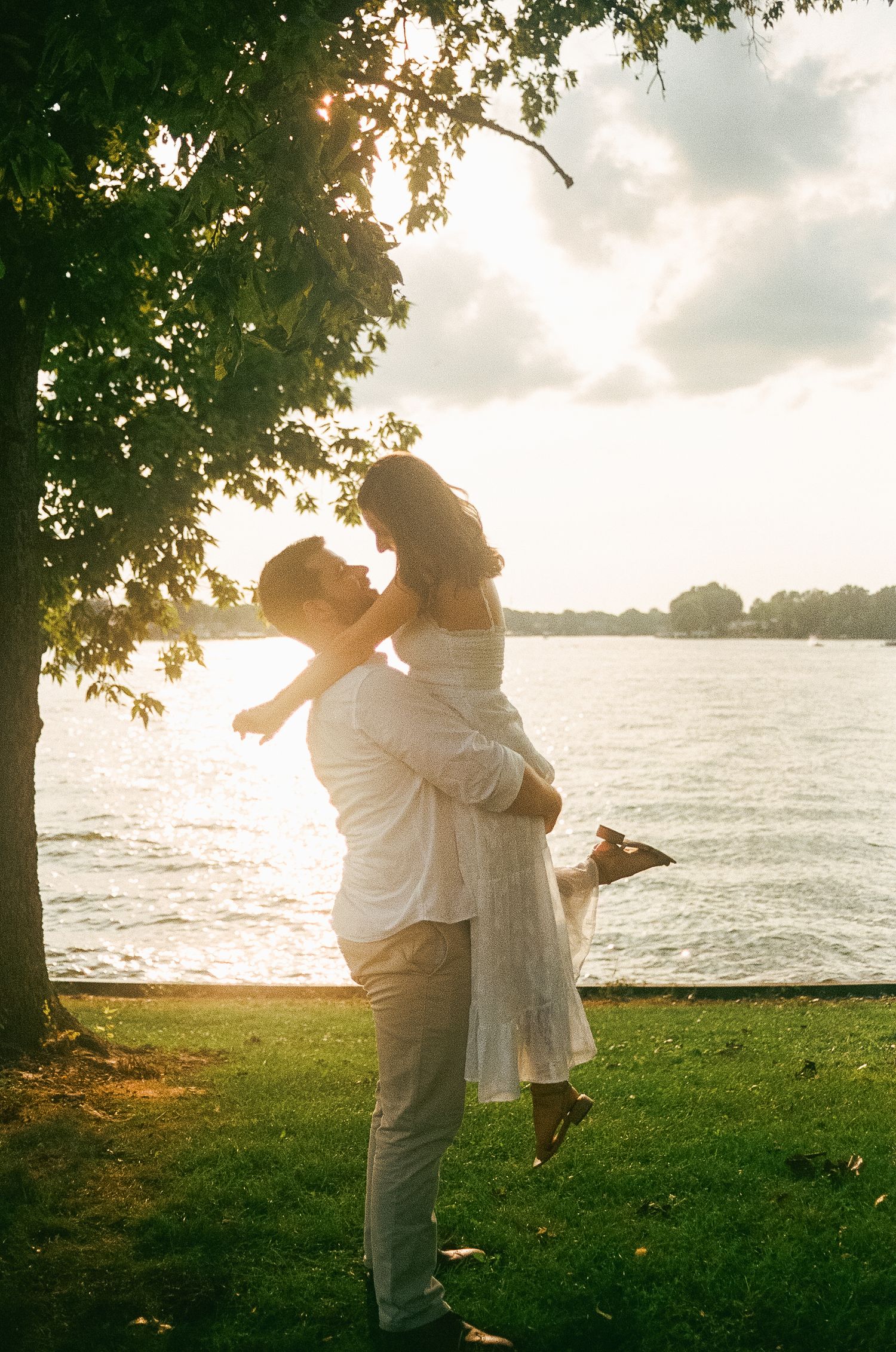 Couple sharing a romantic moment at sunset by a lake, man lifting woman in white dress under a tree.