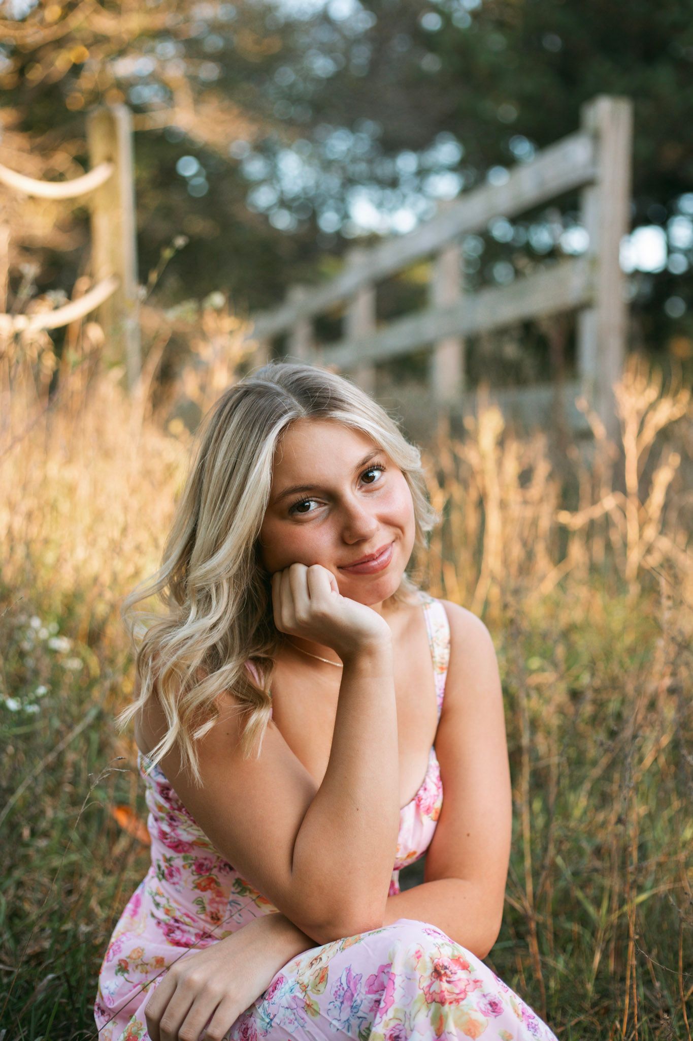 Smiling blonde teen girl in floral dress sitting in golden sunlit field near rustic wooden fence.