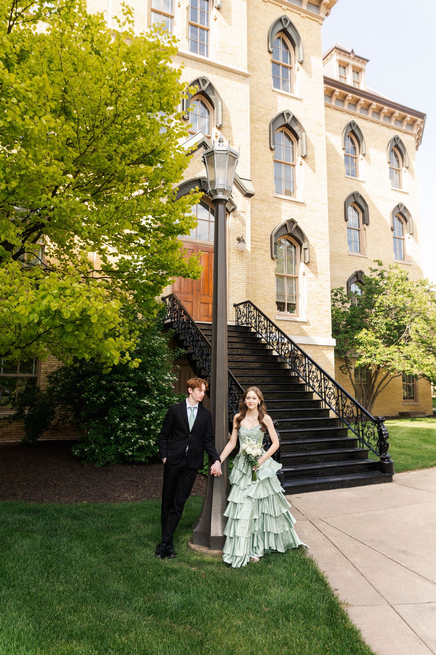 A couple in formal attire walks hand in hand outside a Gothic-style building in spring.