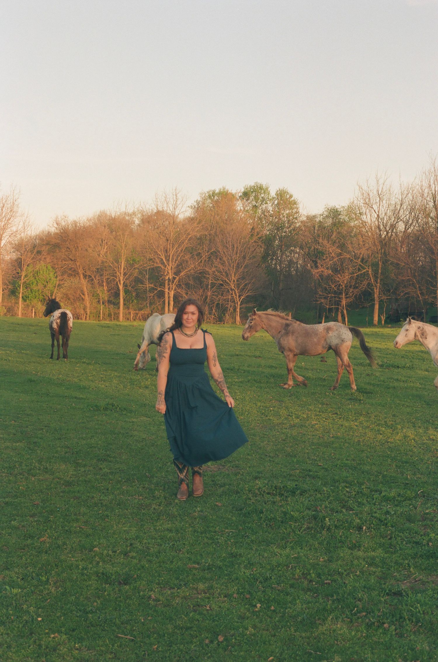 Woman in teal dress stands confidently as horses roam a lush green pasture at sunset.