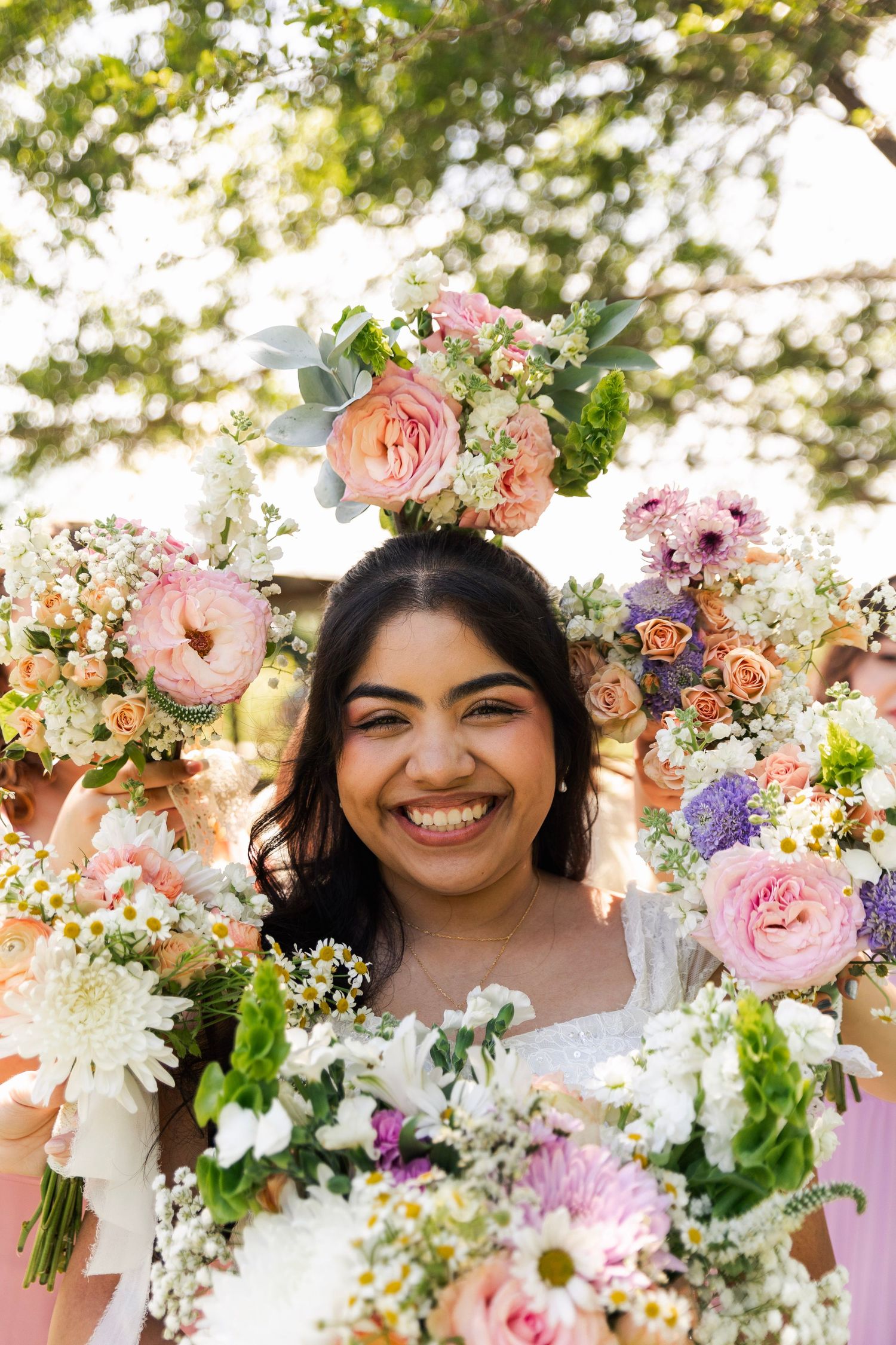 A joyful bride smiles broadly surrounded by colorful pink and white wedding bouquets held by bridesmaids outdoors.