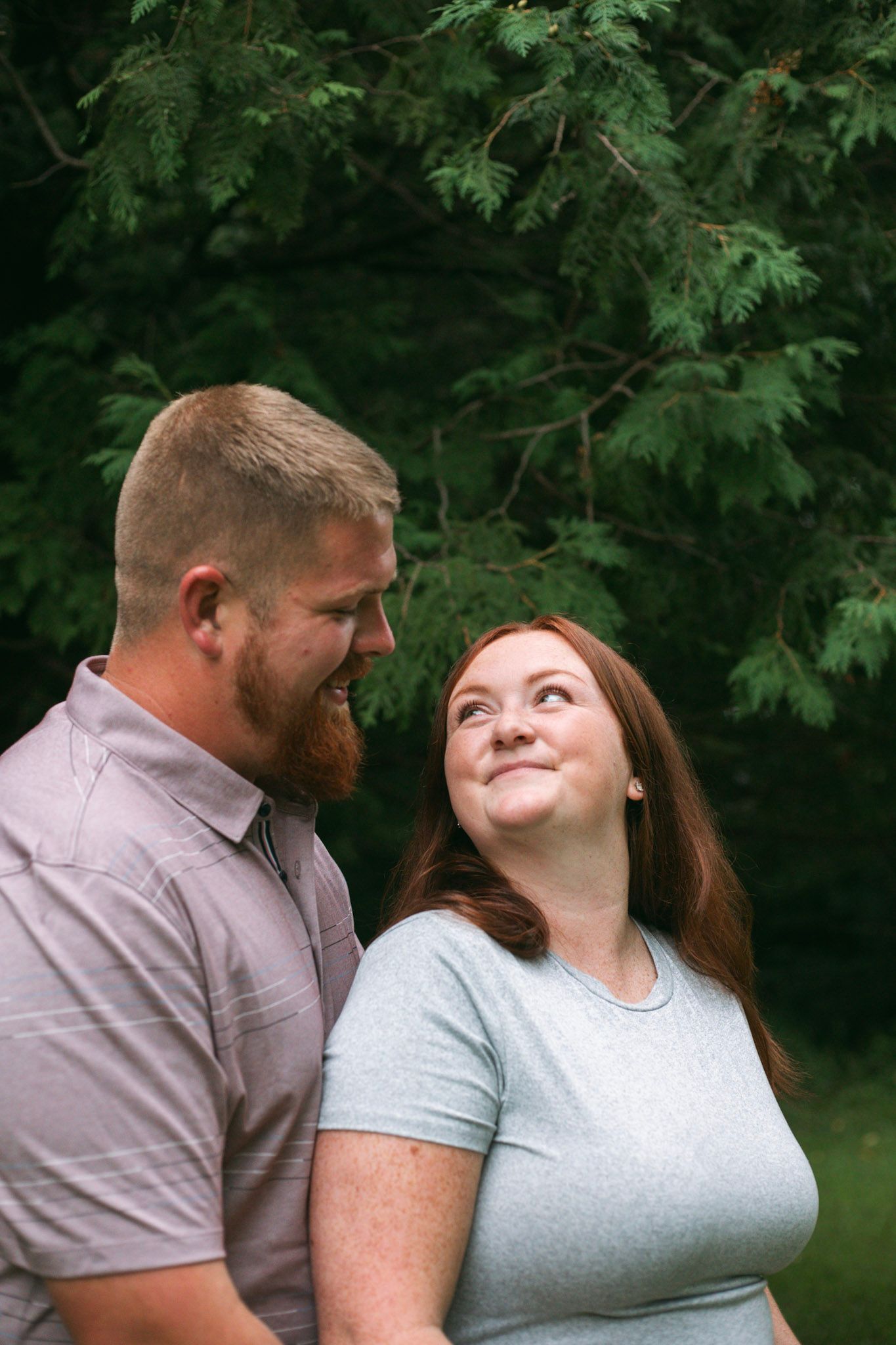 Smiling couple looking at each other affectionately beneath green tree branches outdoors.