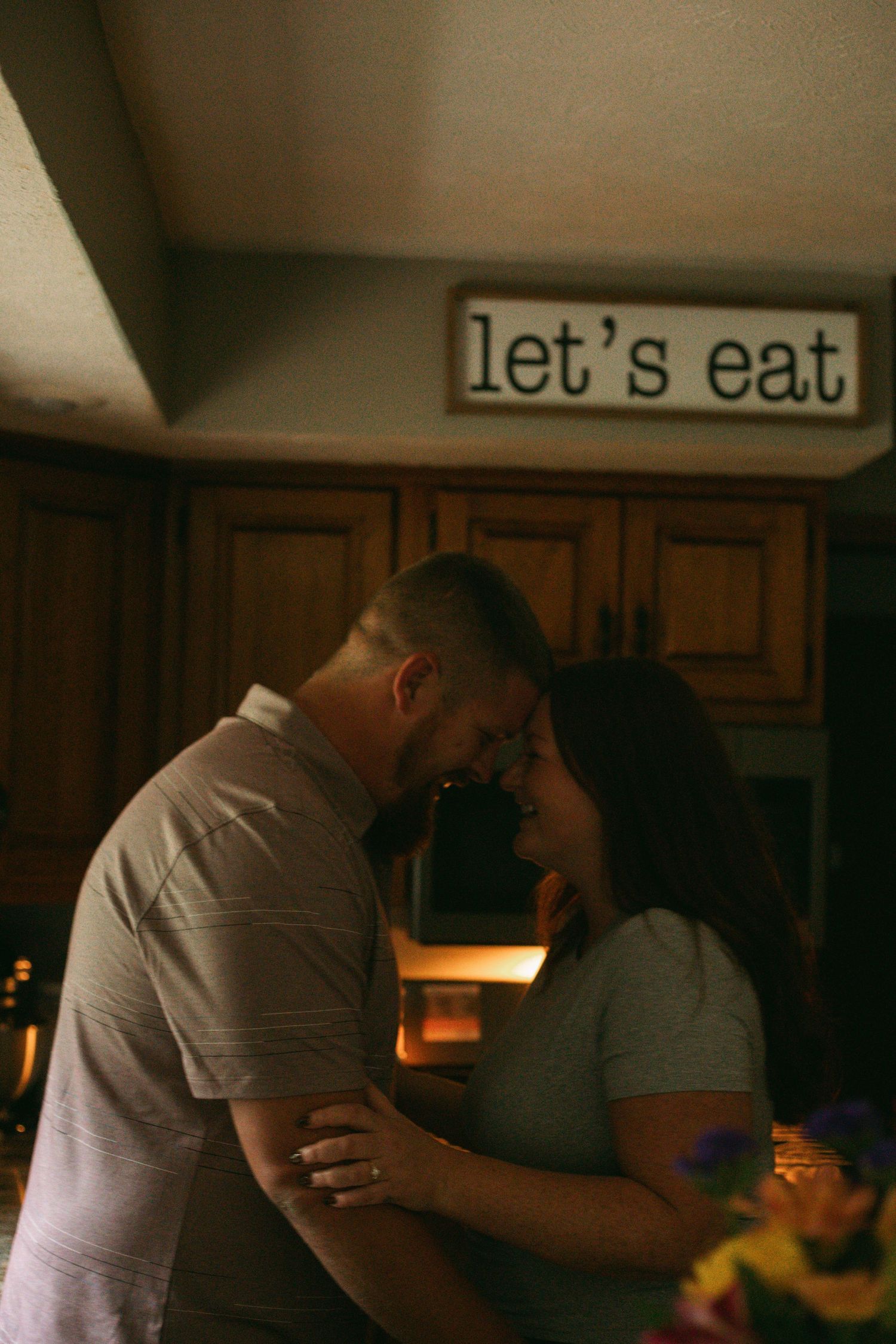 A couple shares an intimate moment in a warmly lit kitchen beneath a rustic let's eat sign.