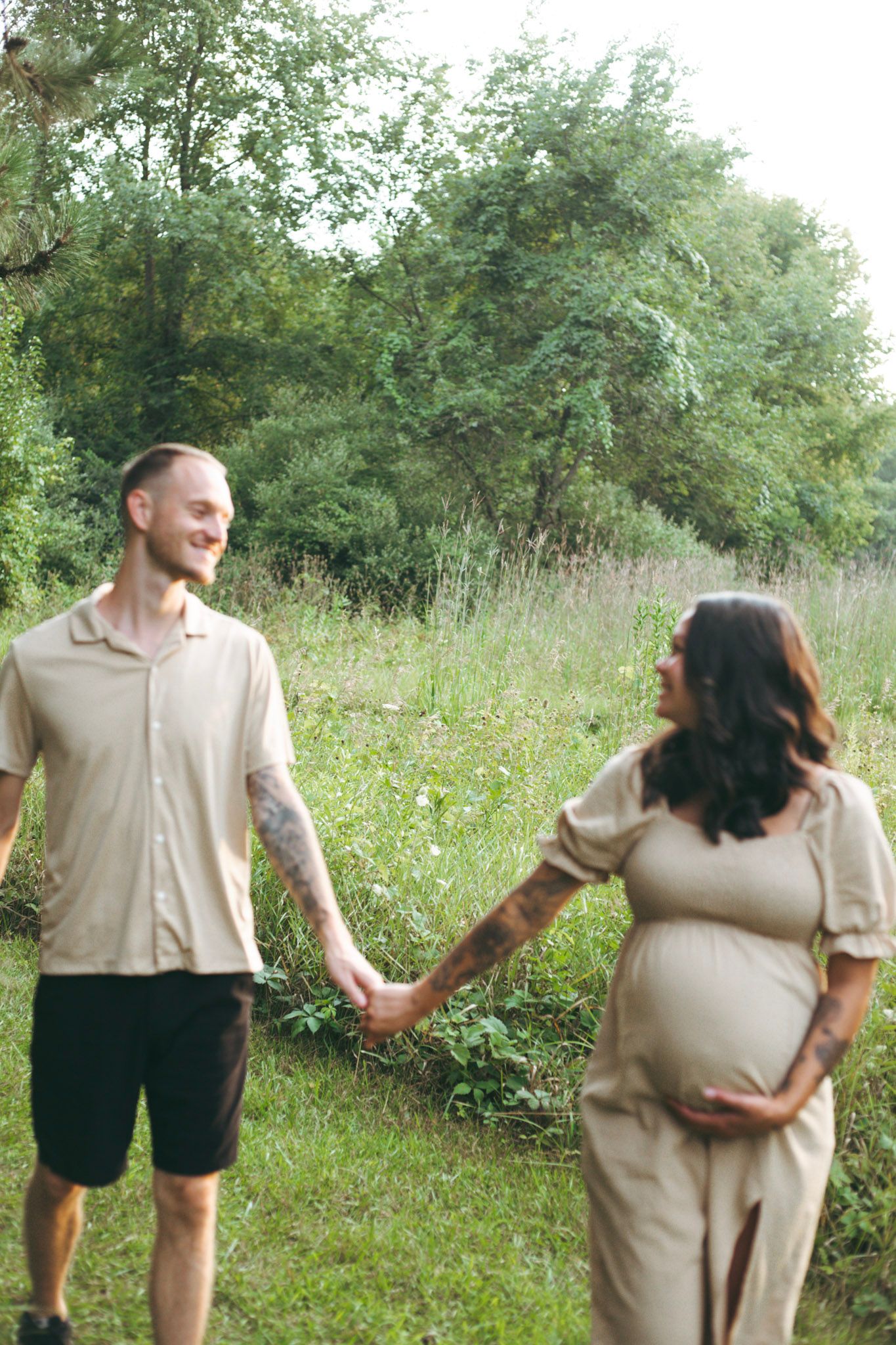 A smiling couple holds hands outdoors, the woman cradling her pregnant belly in a lush green setting.